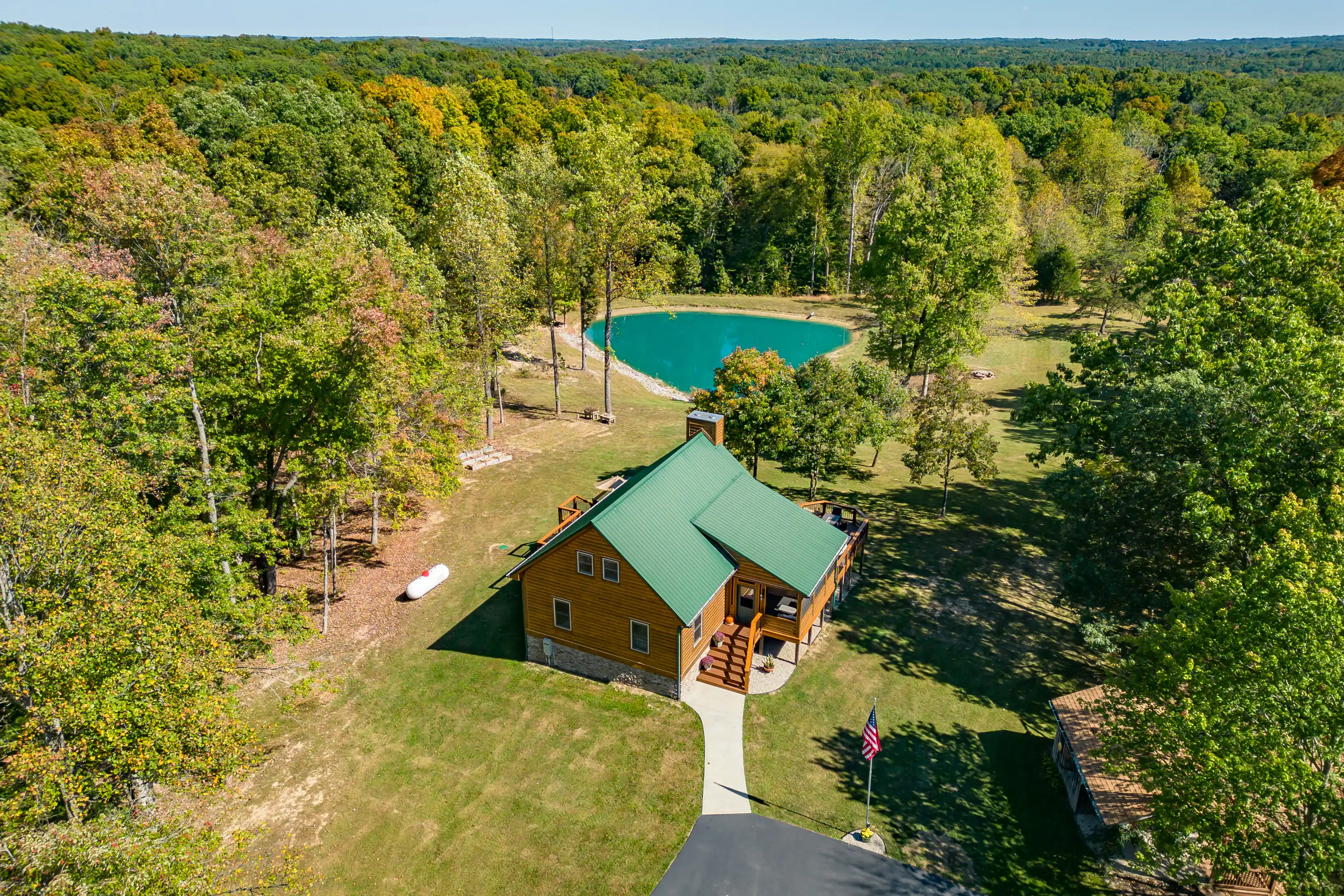 Aerial view of a log cabin with a green roof surrounded by lush trees, next to a small pond in a forested area, with an American flag in the foreground.