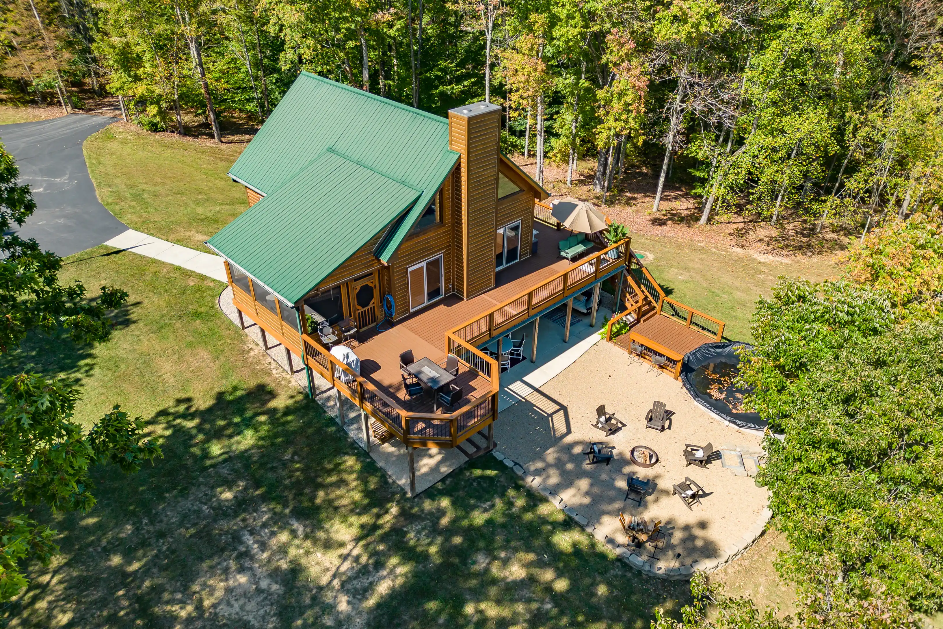 Aerial view of a two-story cabin with a green roof and large deck, surrounded by trees with a fire pit area and trampoline in the backyard.