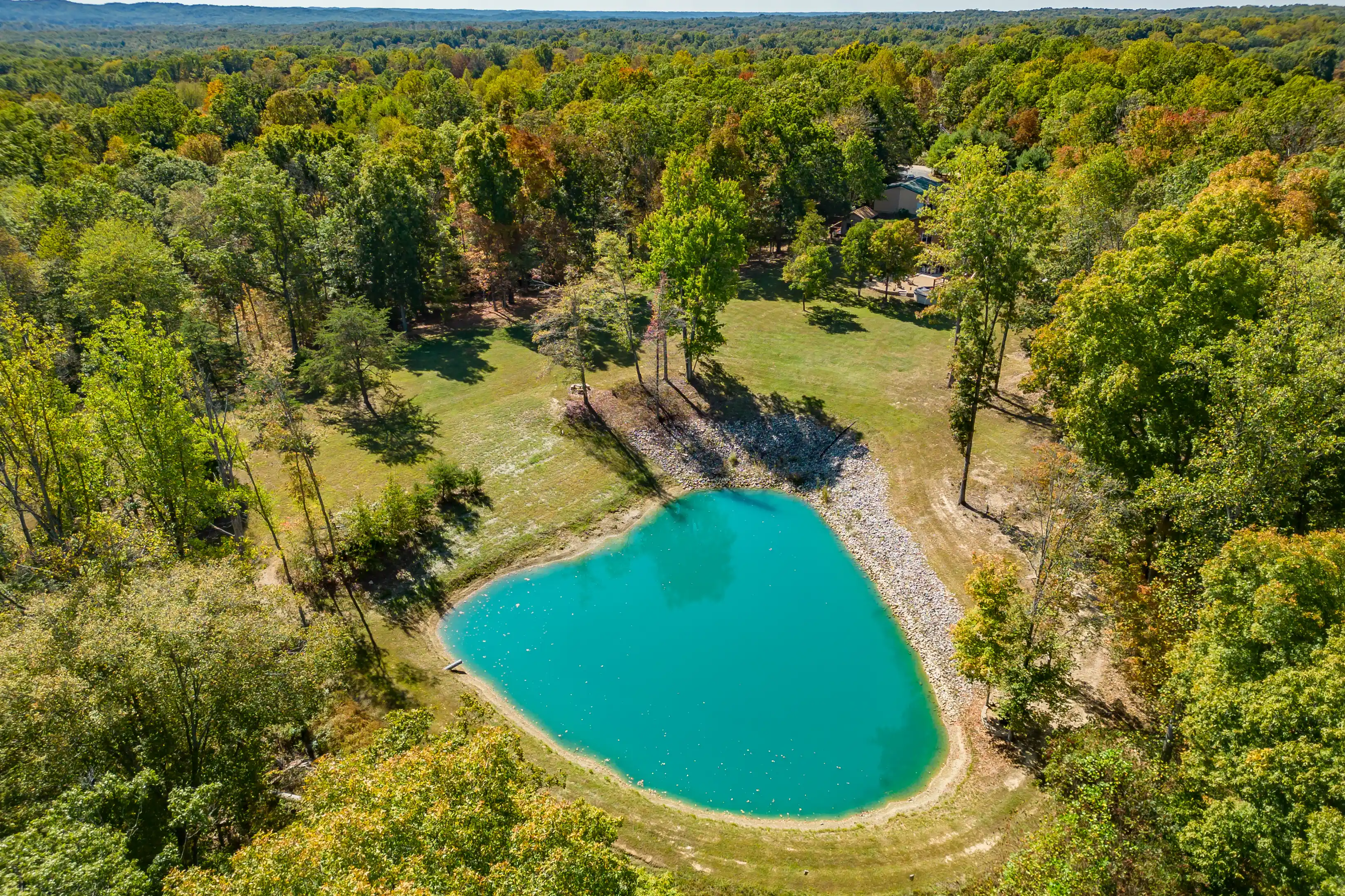 Aerial view of a small heart-shaped pond surrounded by trees with fall foliage and a house partially visible in the background.