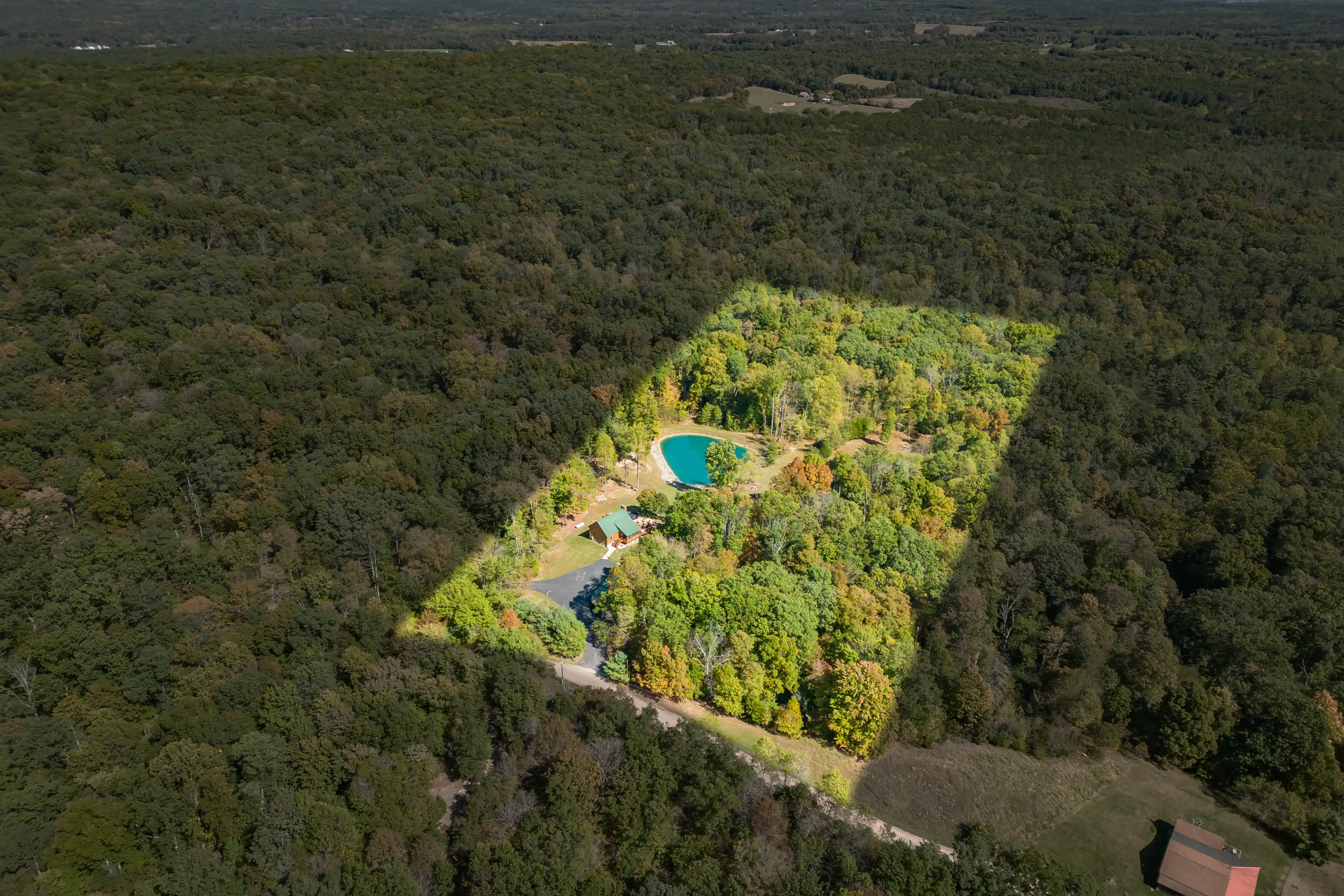 Aerial view of a kidney-shaped swimming pool amidst a forest clearing with a building close by and vast forest extending into the horizon.