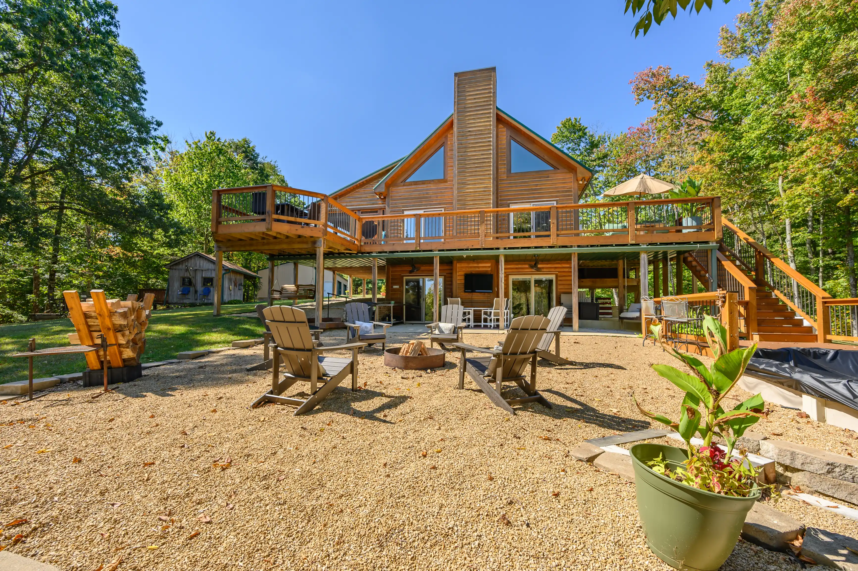 A sunny view of a large wooden cabin with a spacious deck and outdoor seating area surrounded by trees.