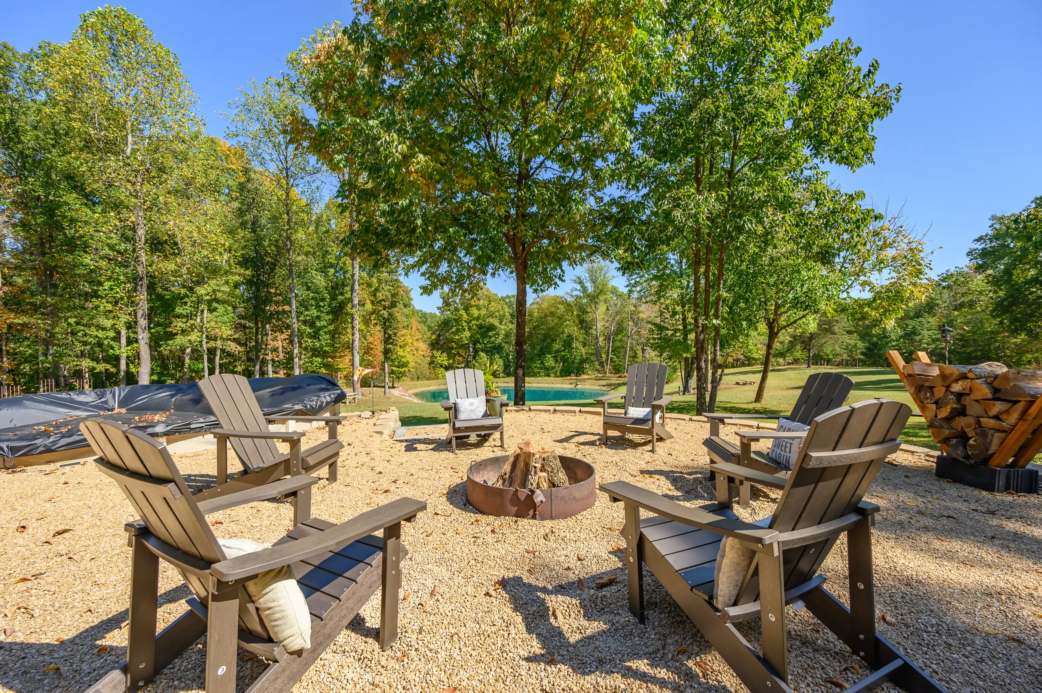 Outdoor fire pit area with Adirondack chairs surrounded by trees on a bed of gravel with a pile of firewood to the side.