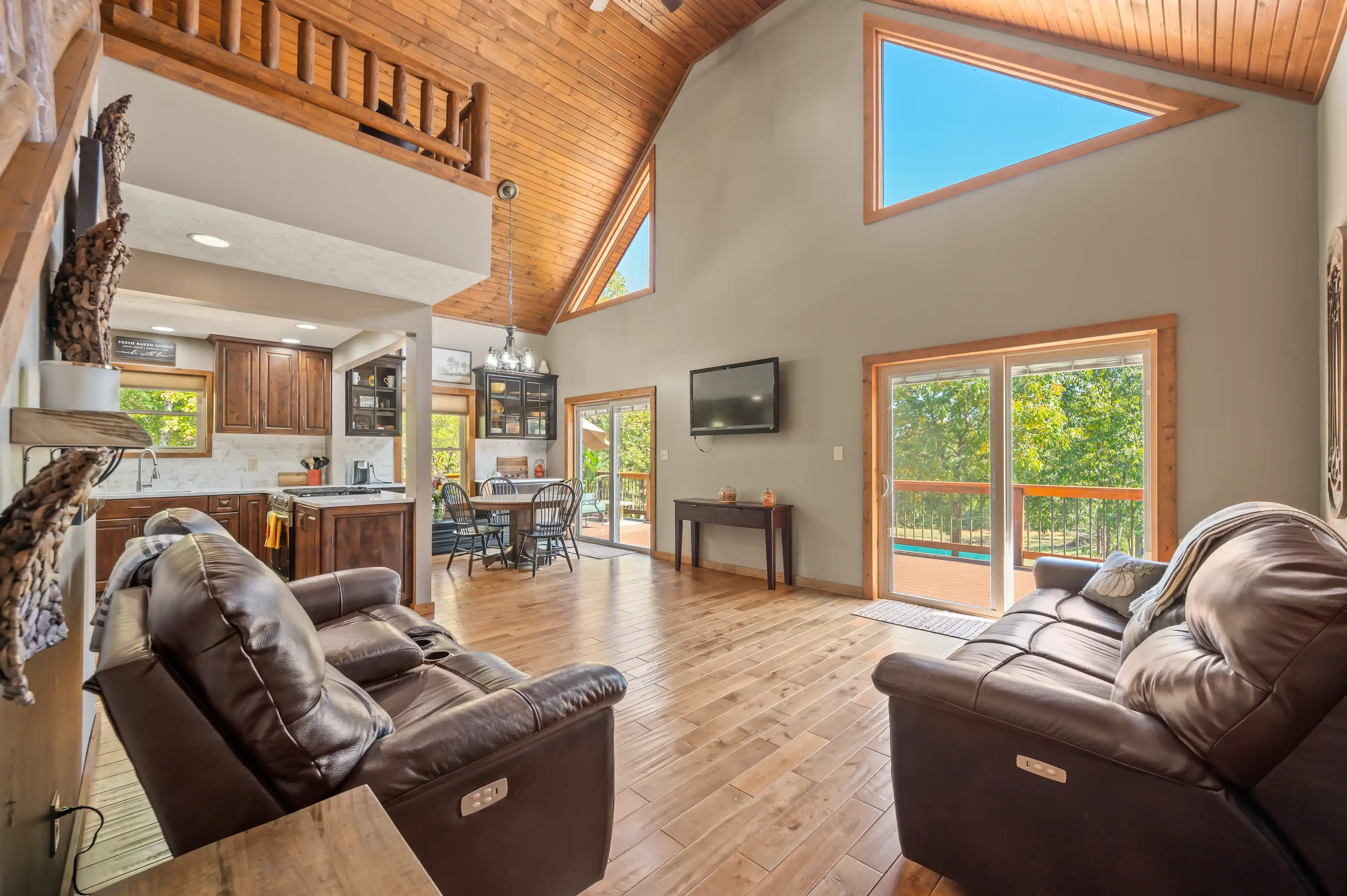 Spacious living room with high ceilings, wooden floors, and a loft, featuring leather sofas, a mounted TV, and sliding doors leading to a balcony with forest views.