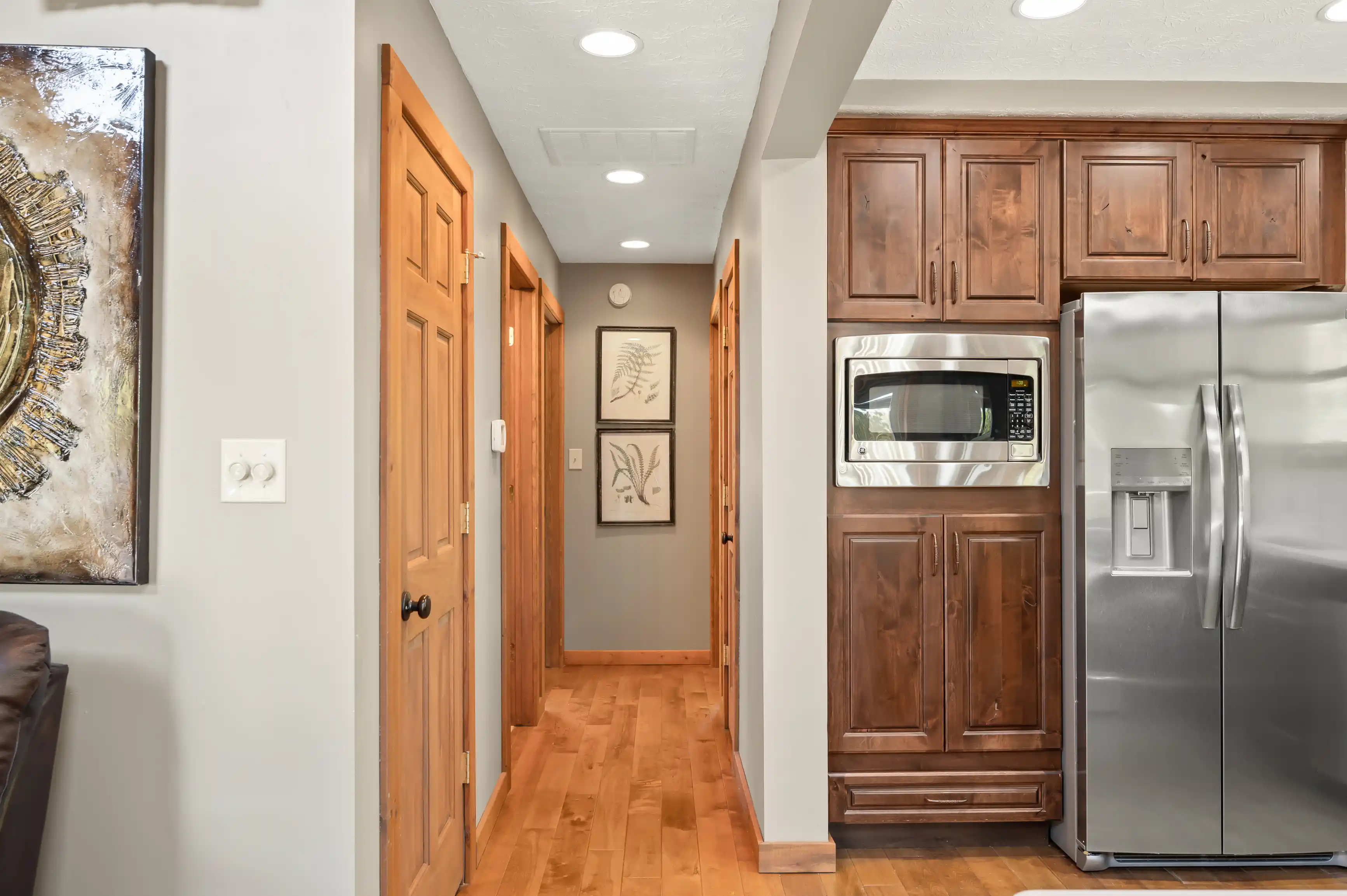 Interior view of a modern kitchen with stainless steel appliances, wooden cabinetry, and a hallway leading to other rooms, featuring hardwood floors and decorated with wall art.