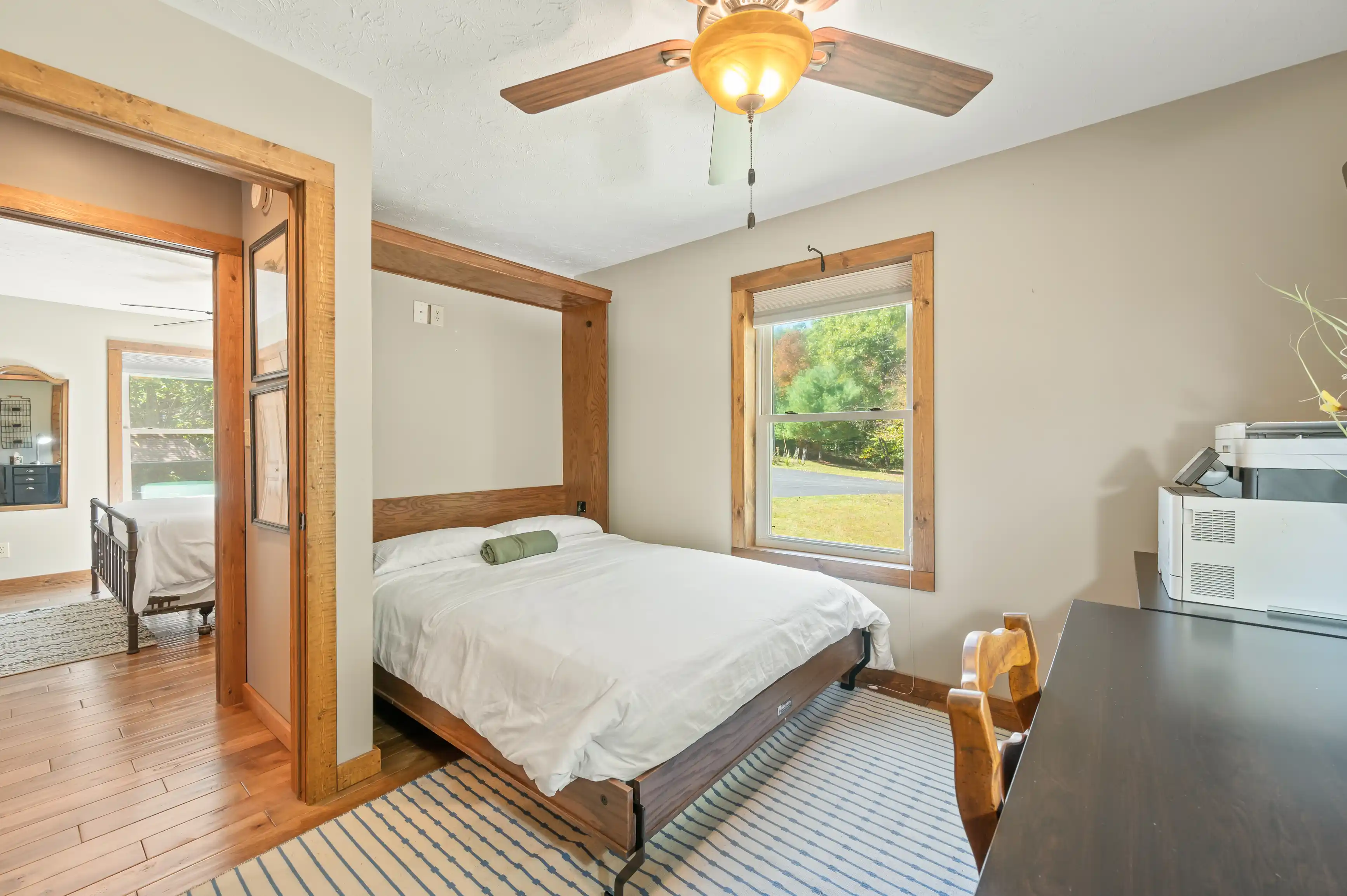 Bright and airy bedroom with a double bed, wooden ceiling fan, floor-to-ceiling wood trim, striped area rug, and a view of the yard through two large windows.