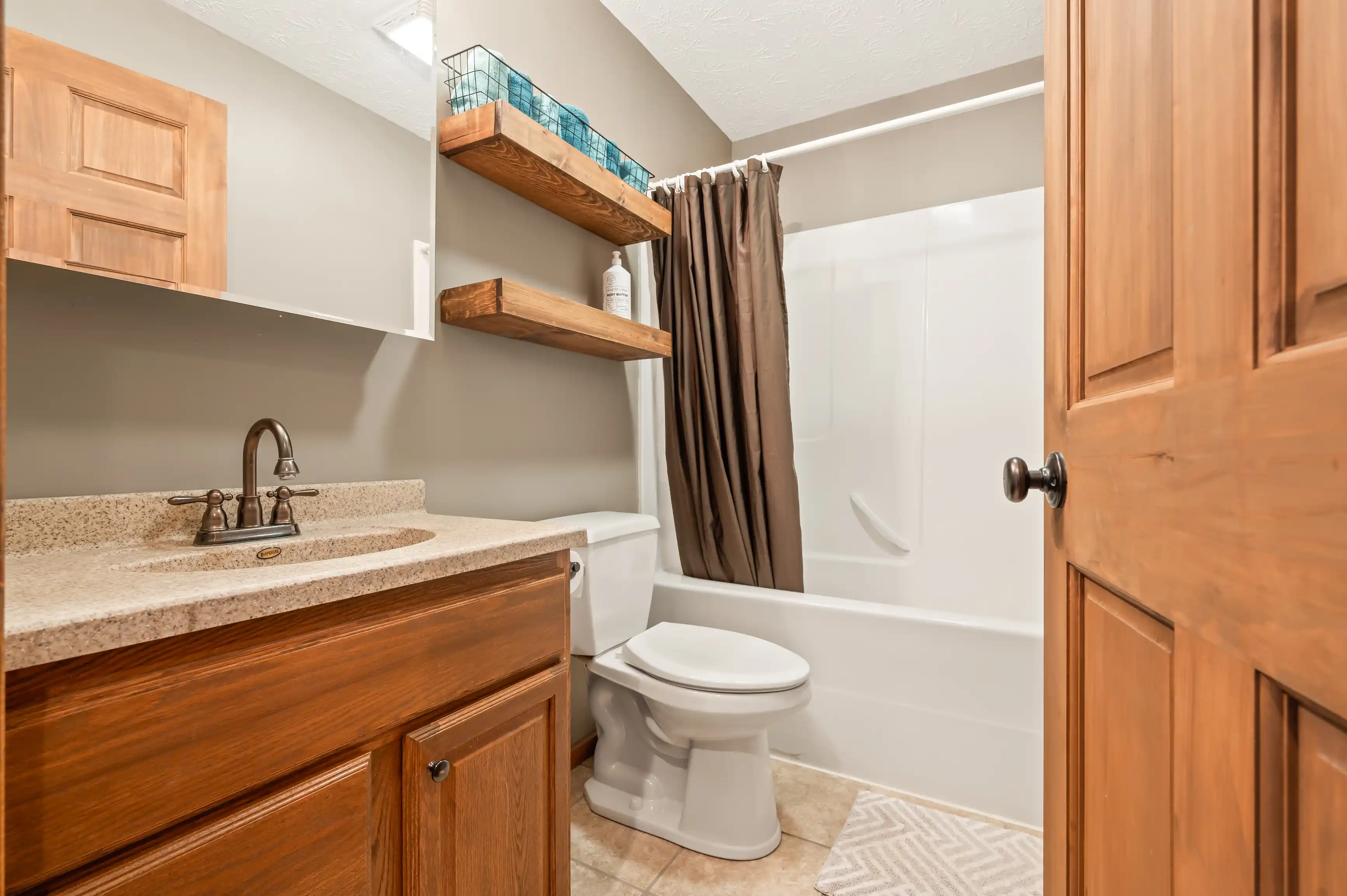 A modern bathroom with wooden cabinetry, granite countertop with undermount sink, two floating shelves above the toilet, and a shower-tub combo with a brown curtain.