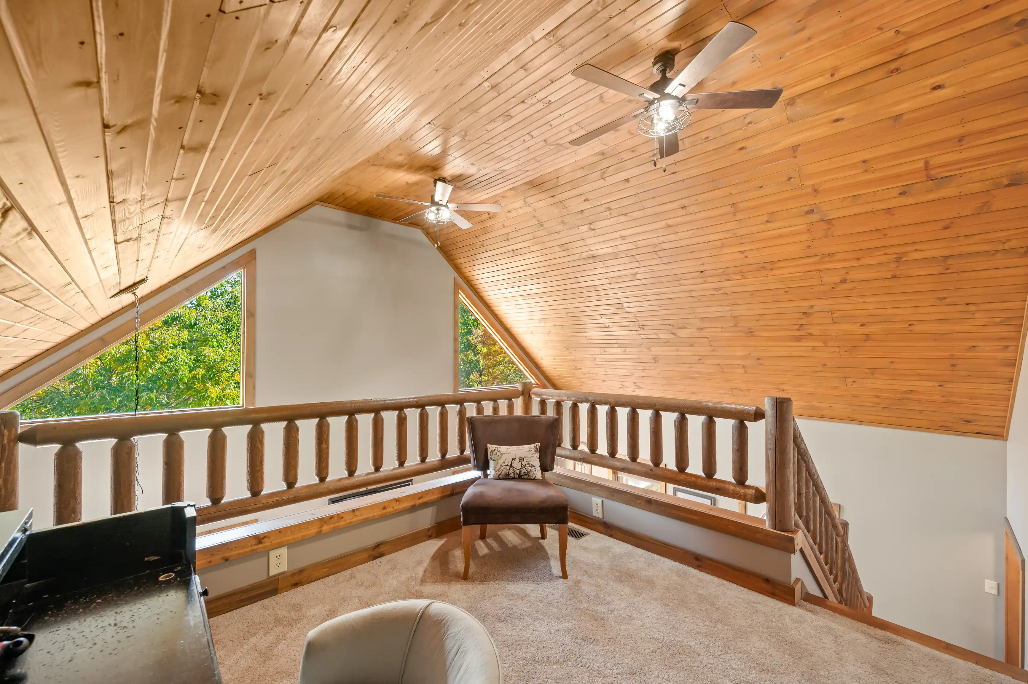Cozy attic loft area with wooden ceiling and flooring, furnished with a brown armchair, featuring large windows with a view of greenery, and two ceiling fans.