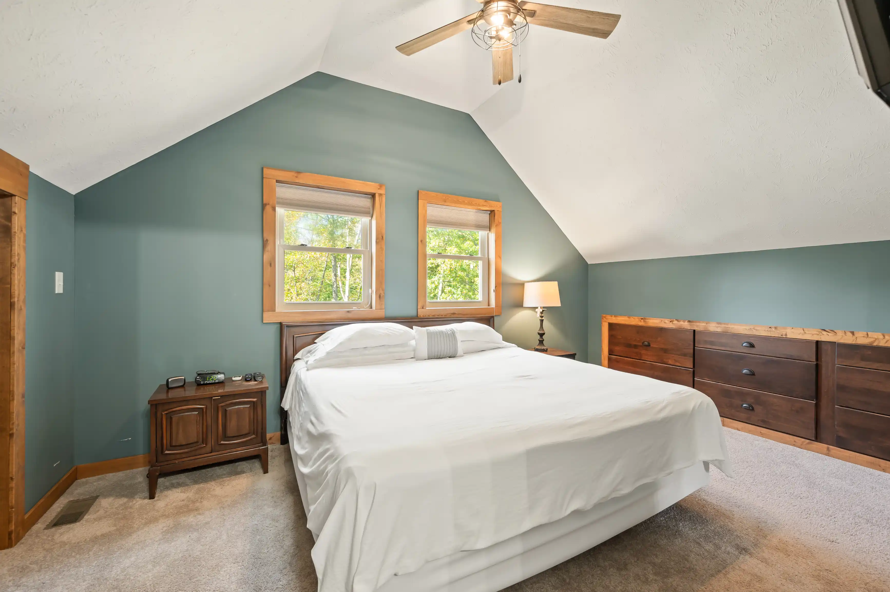 Cozy bedroom with slanted ceiling, teal accent wall, large bed with white bedding, wooden furniture, and a ceiling fan. Two windows show greenery outside.