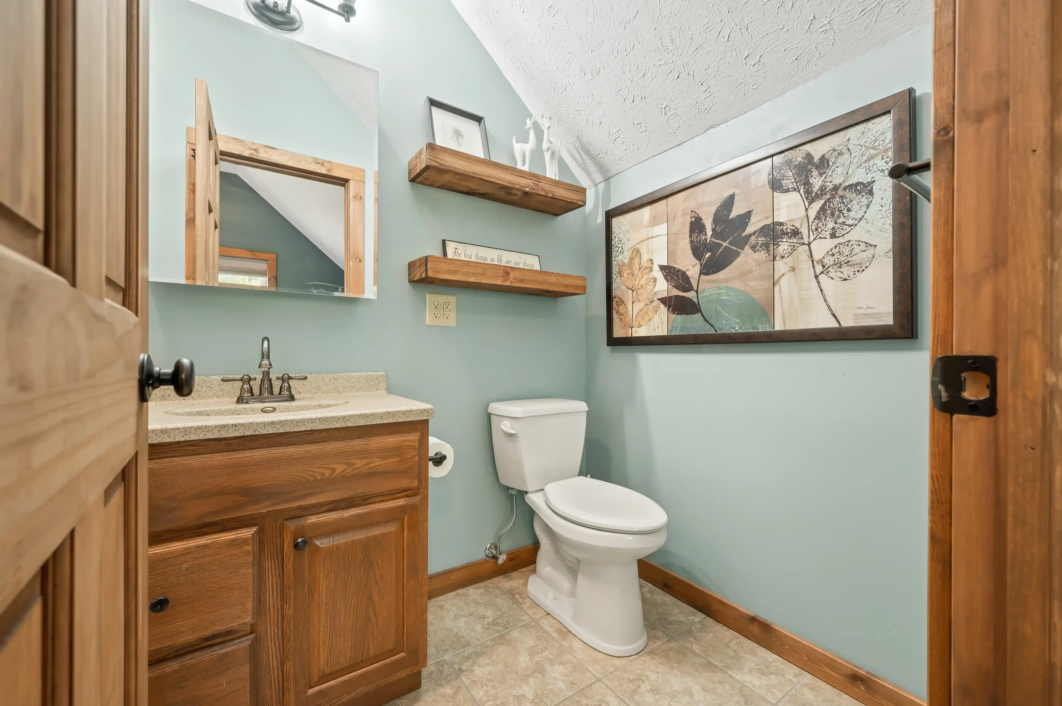 A cozy bathroom interior with teal walls, featuring a wooden vanity with a granite countertop and a mirror above, a toilet, decorative floating shelves, and a framed botanical print.