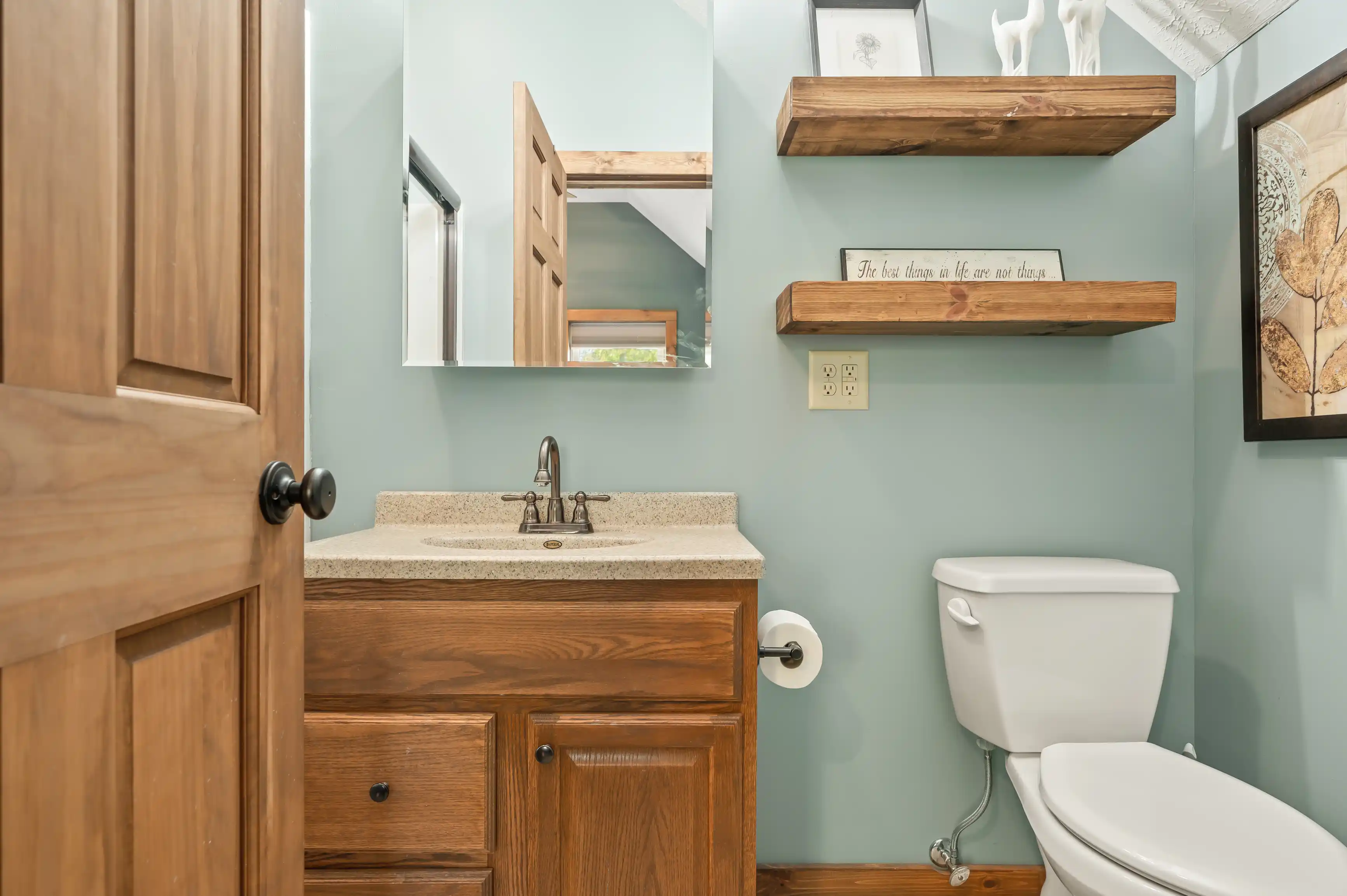 A small bathroom with aqua-colored walls, wooden vanity with granite countertop, mirror, floating wooden shelves, toilet, and wall art.