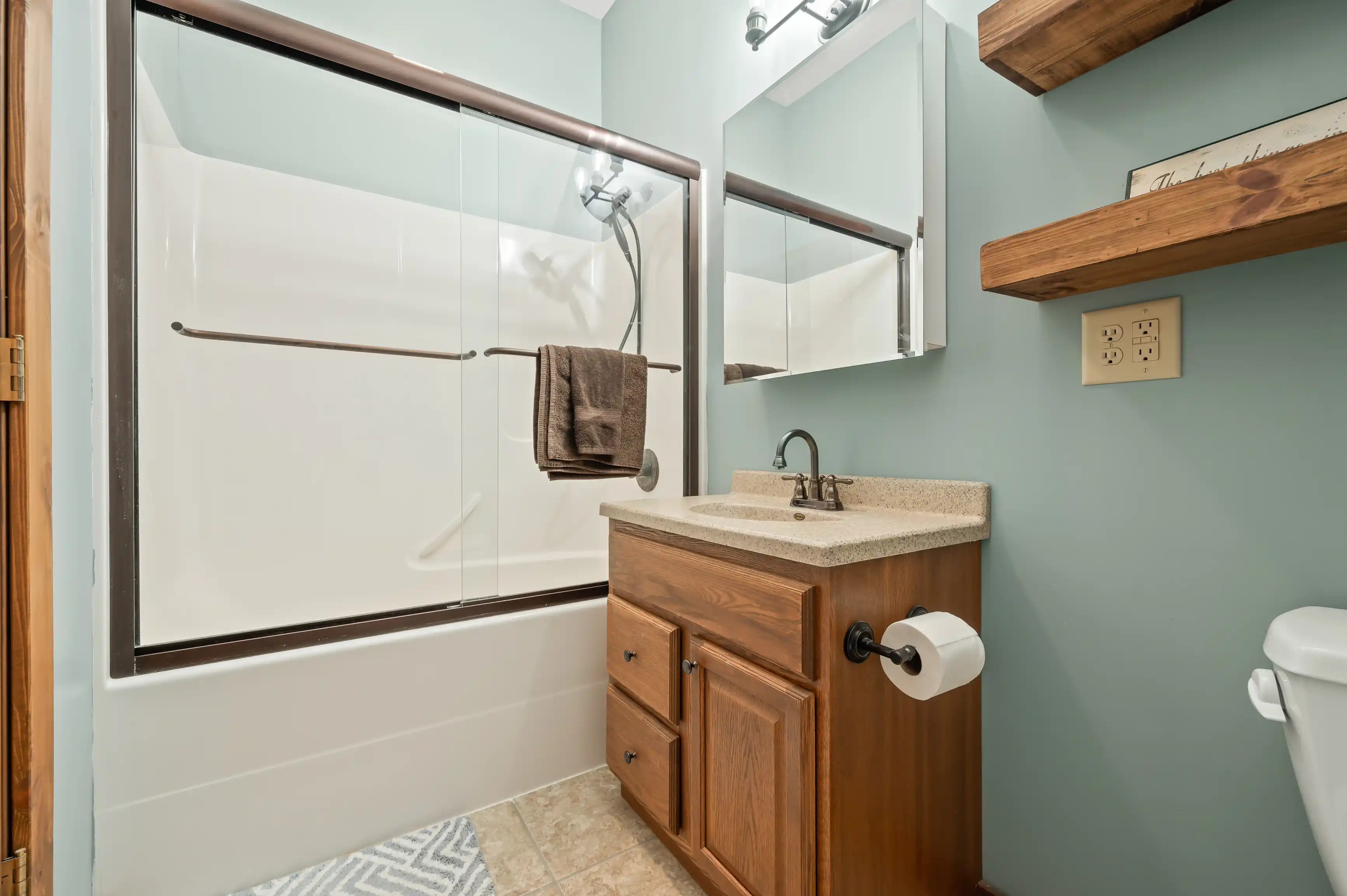 A modern bathroom with a sliding glass door shower, wooden vanity with a granite top, large mirror, floating wooden shelves, and a toilet.