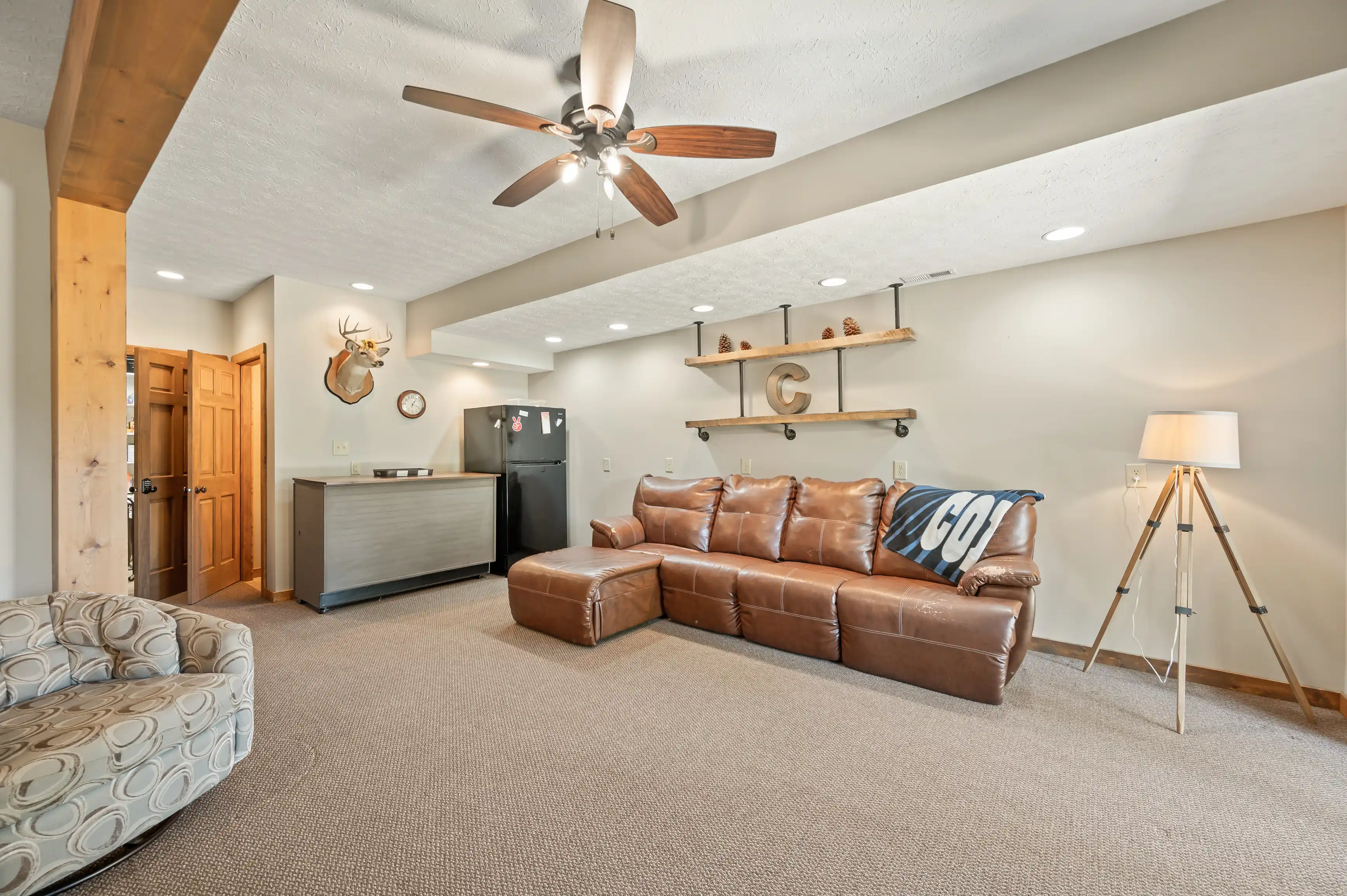 Spacious living room with textured ceiling and fan, leather sectional sofa, tripod floor lamp, floating wooden shelves, deer head wall mount, and a kitchenette with modern appliances in the background.