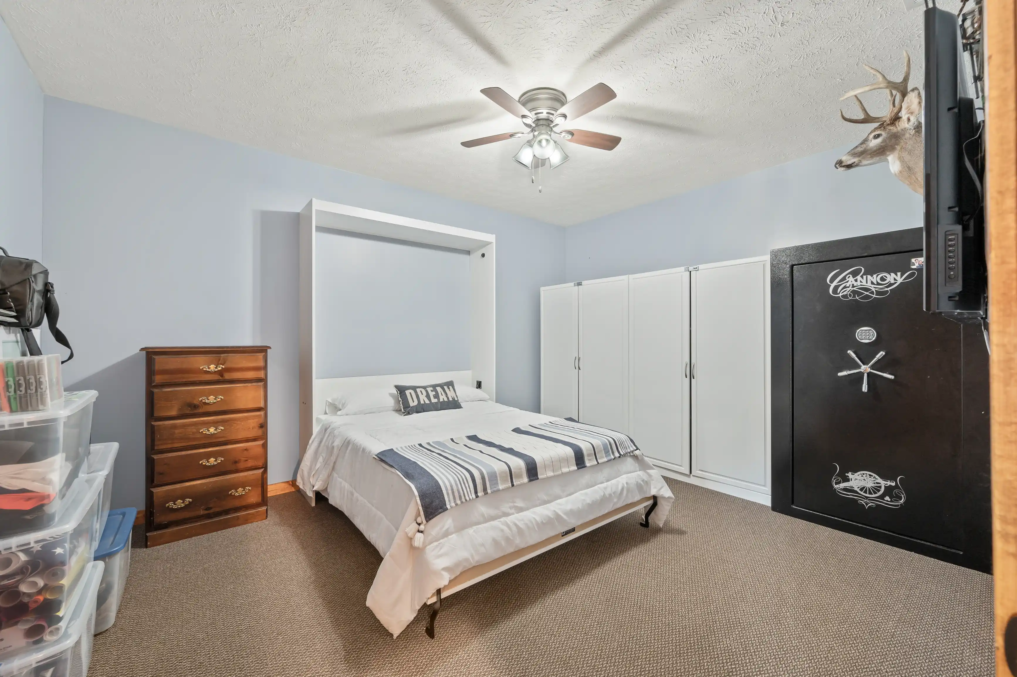 A well-lit bedroom with light blue walls, a white ceiling fan, a double bed with a striped blue-and-white comforter, a brown wooden dresser, white wardrobes, a wall-mounted deer head, and a large black safe with stickers.