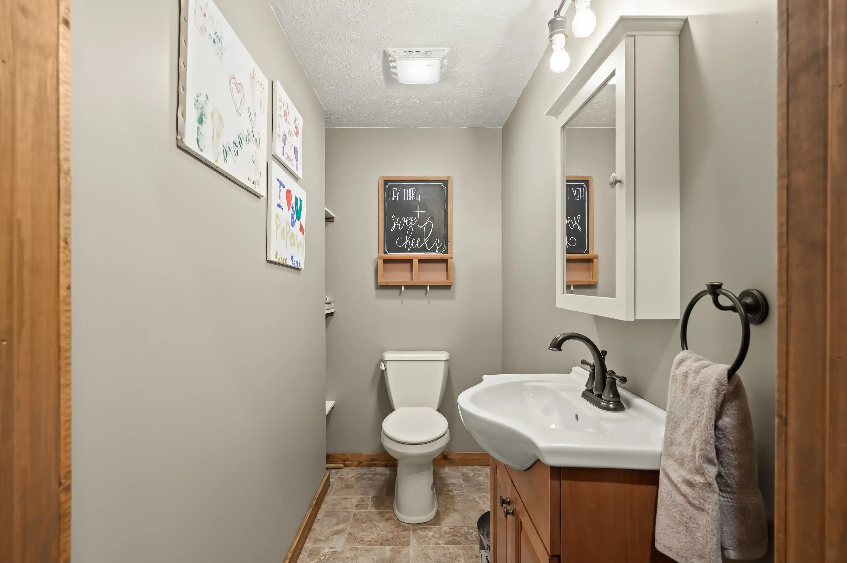 Interior of a cozy bathroom featuring a toilet, a sink with a wooden vanity, a mirror with reflections of a decorative chalkboard, and children's artwork on the wall.