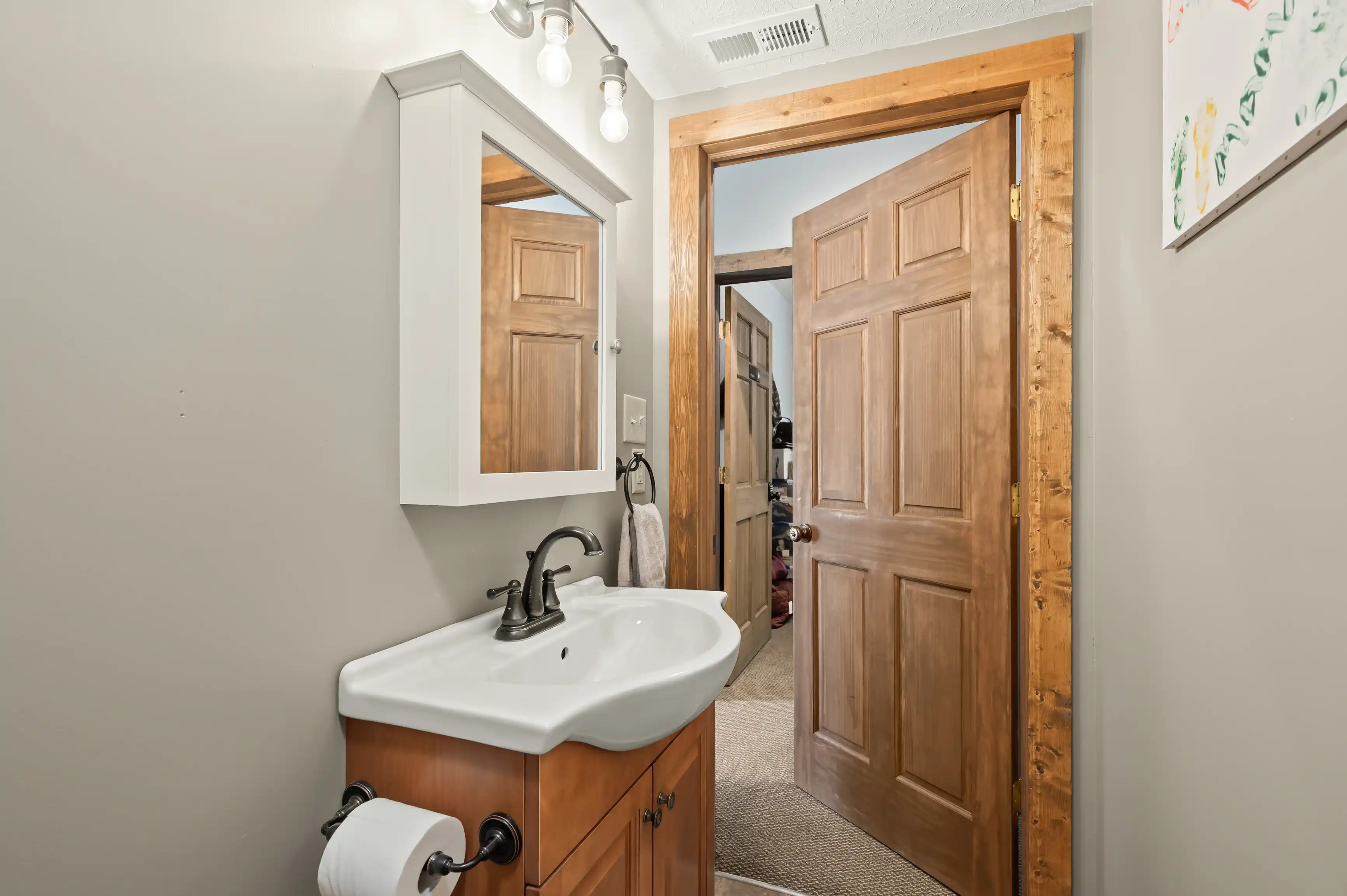 Interior of a well-lit bathroom featuring a wooden cabinet with a sink, a mirrored medicine cabinet above, and an open wooden door leading to another room.