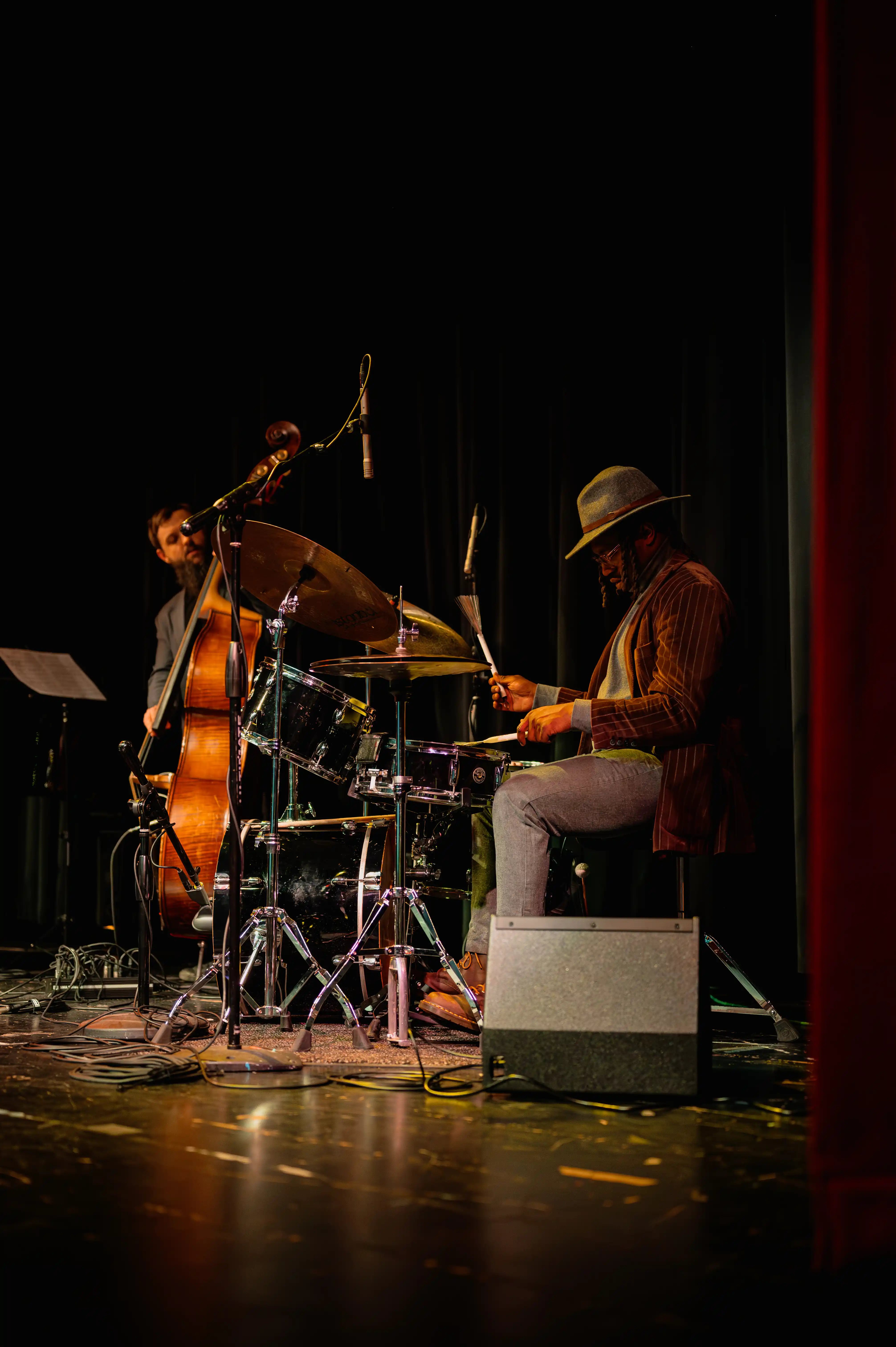 Musicians on stage with a drummer in the foreground and a double bass player in the background, performing under a spotlight.