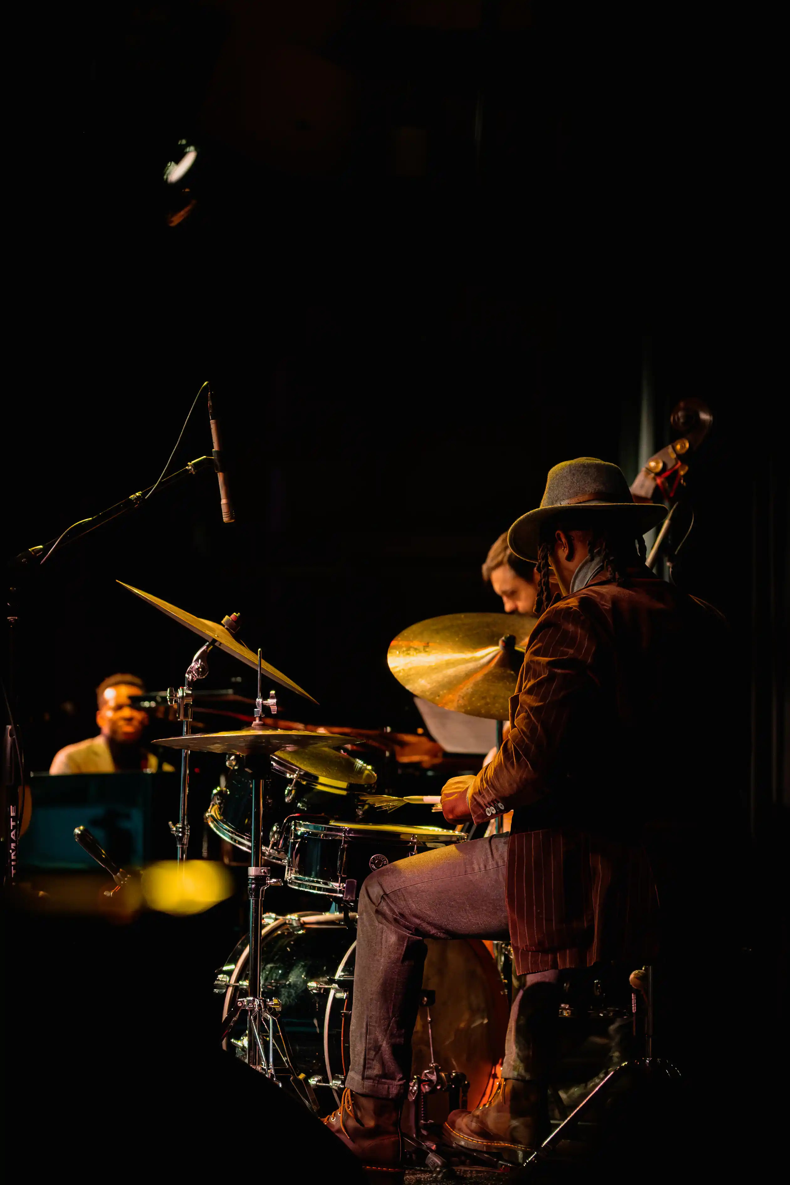 A musician sitting and playing the cello on a dark stage with a drummer in the background.