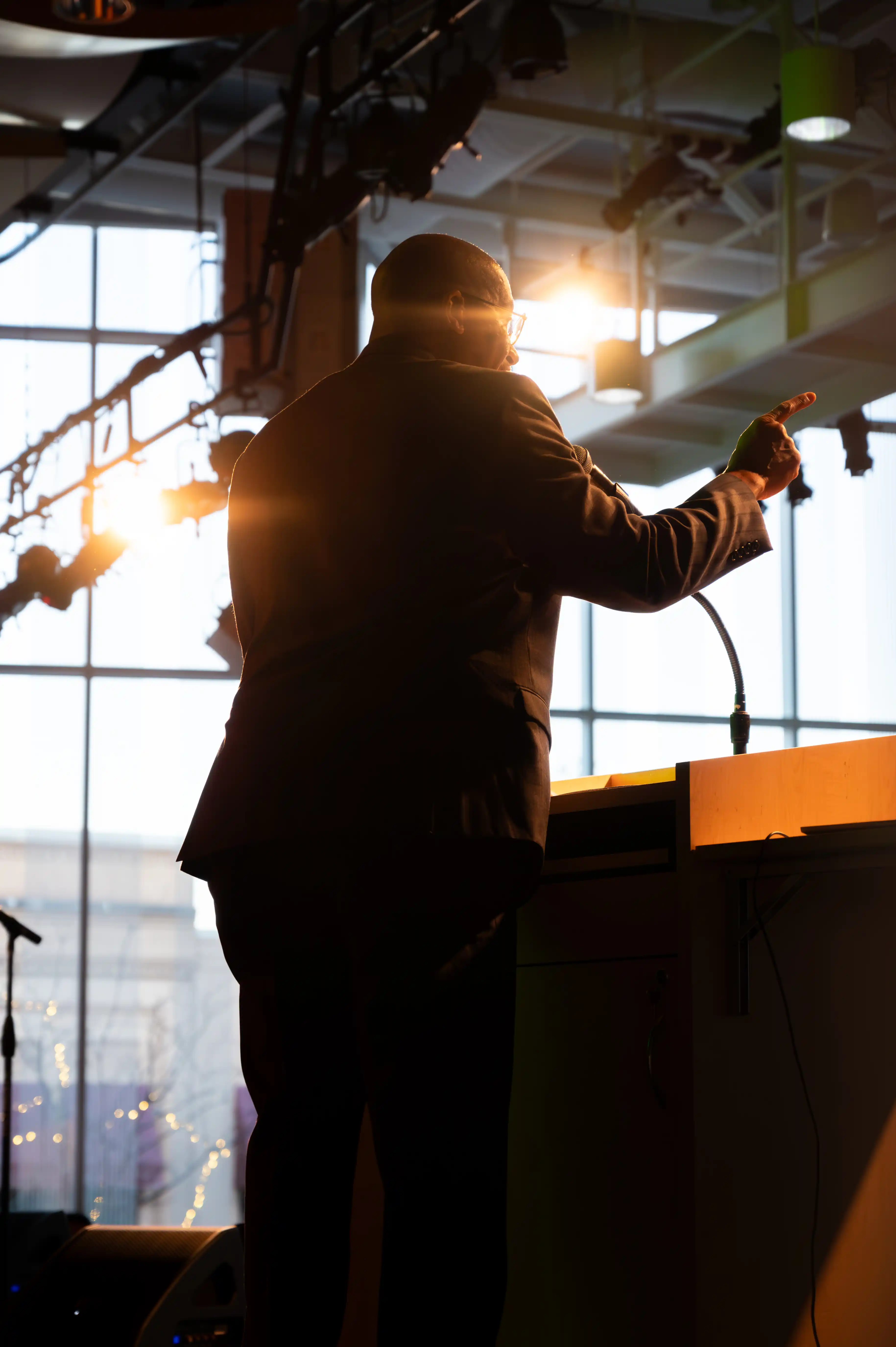 Silhouette of a person speaking at a podium with microphone in a room illuminated by natural light from windows.