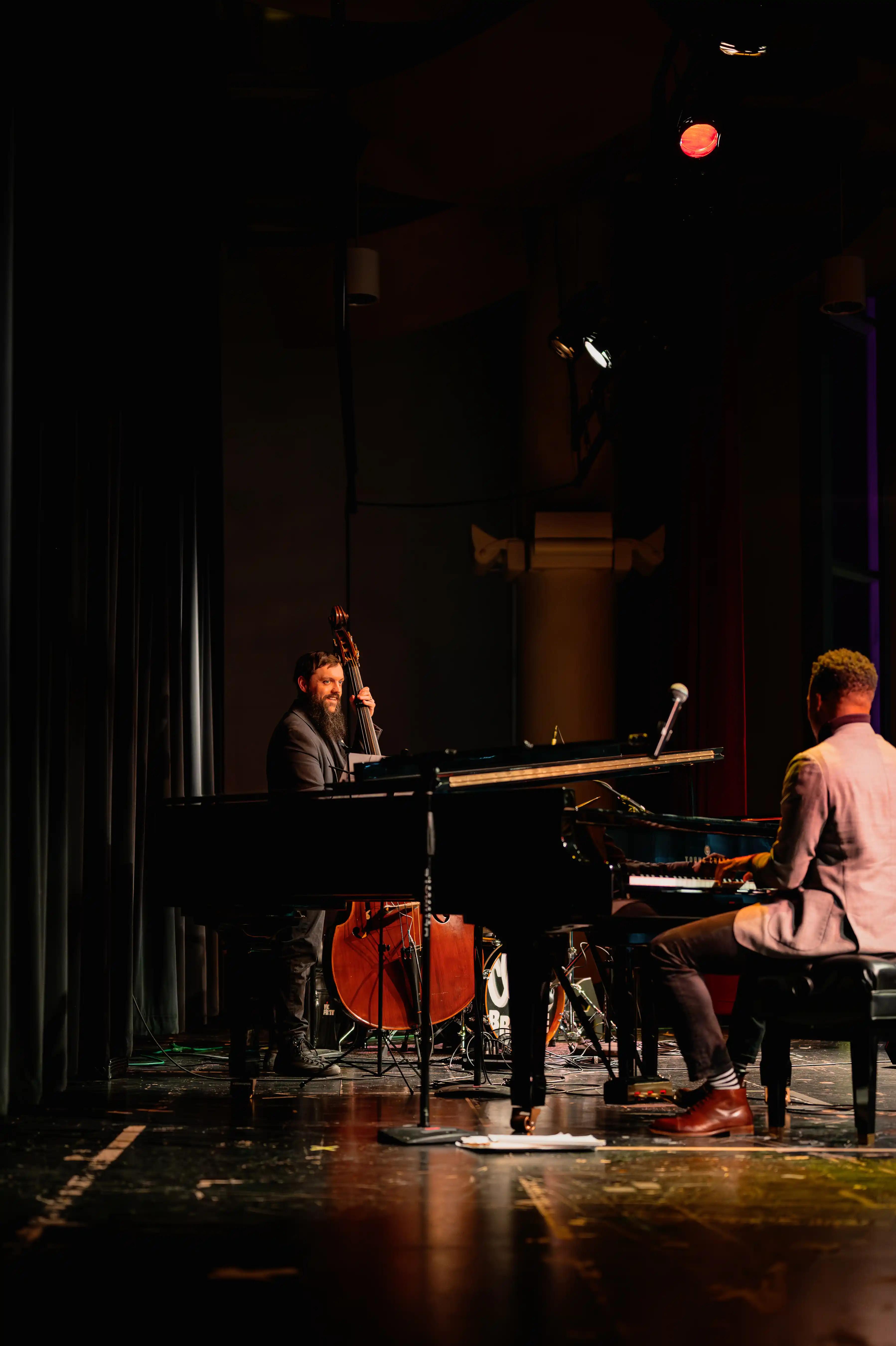 Two musicians on stage with a pianist facing the grand piano and another musician with a saxophone sitting, both under spotlight.