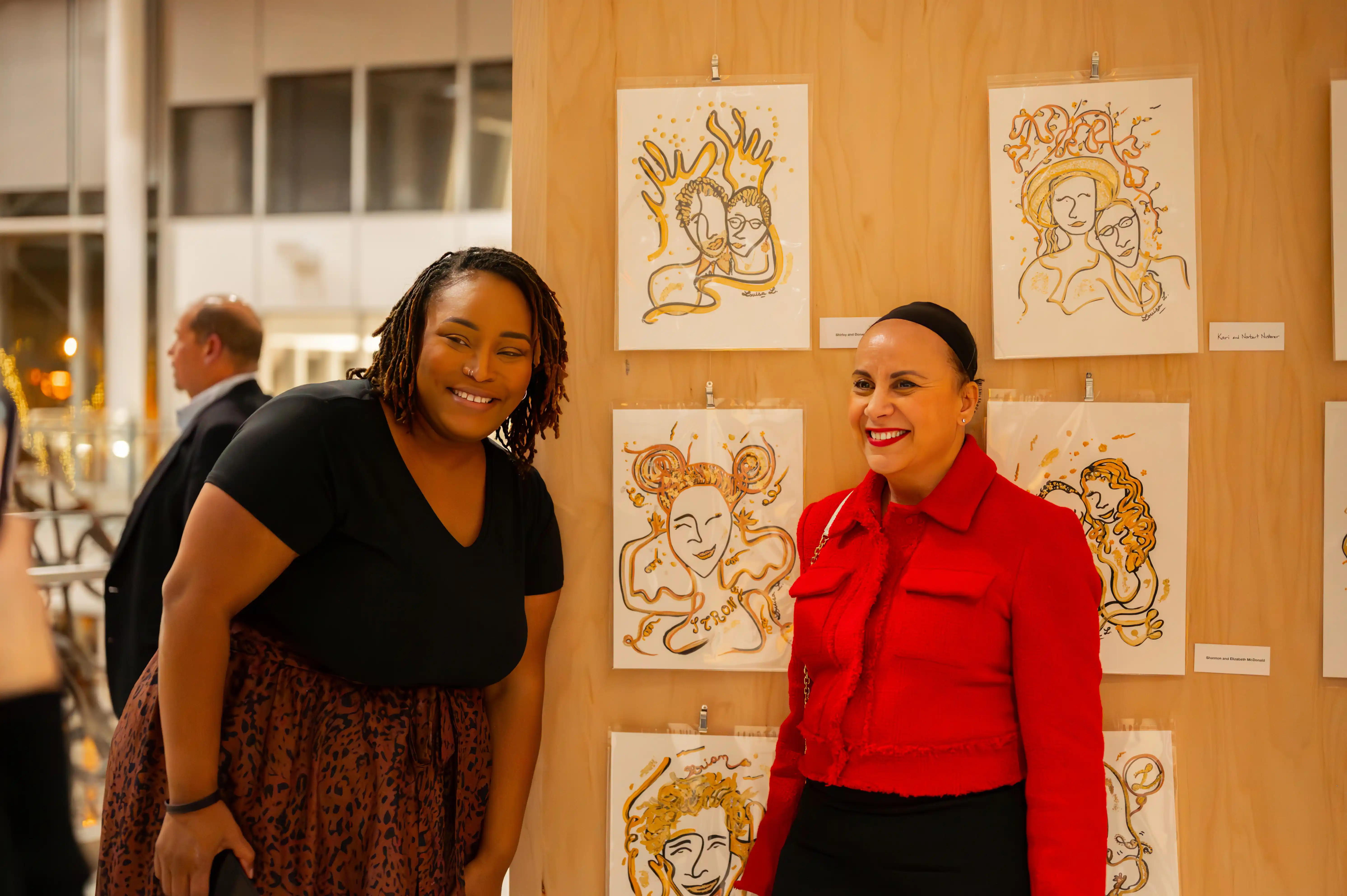 Two women smiling at an art exhibition with drawings displayed on the wall behind them.