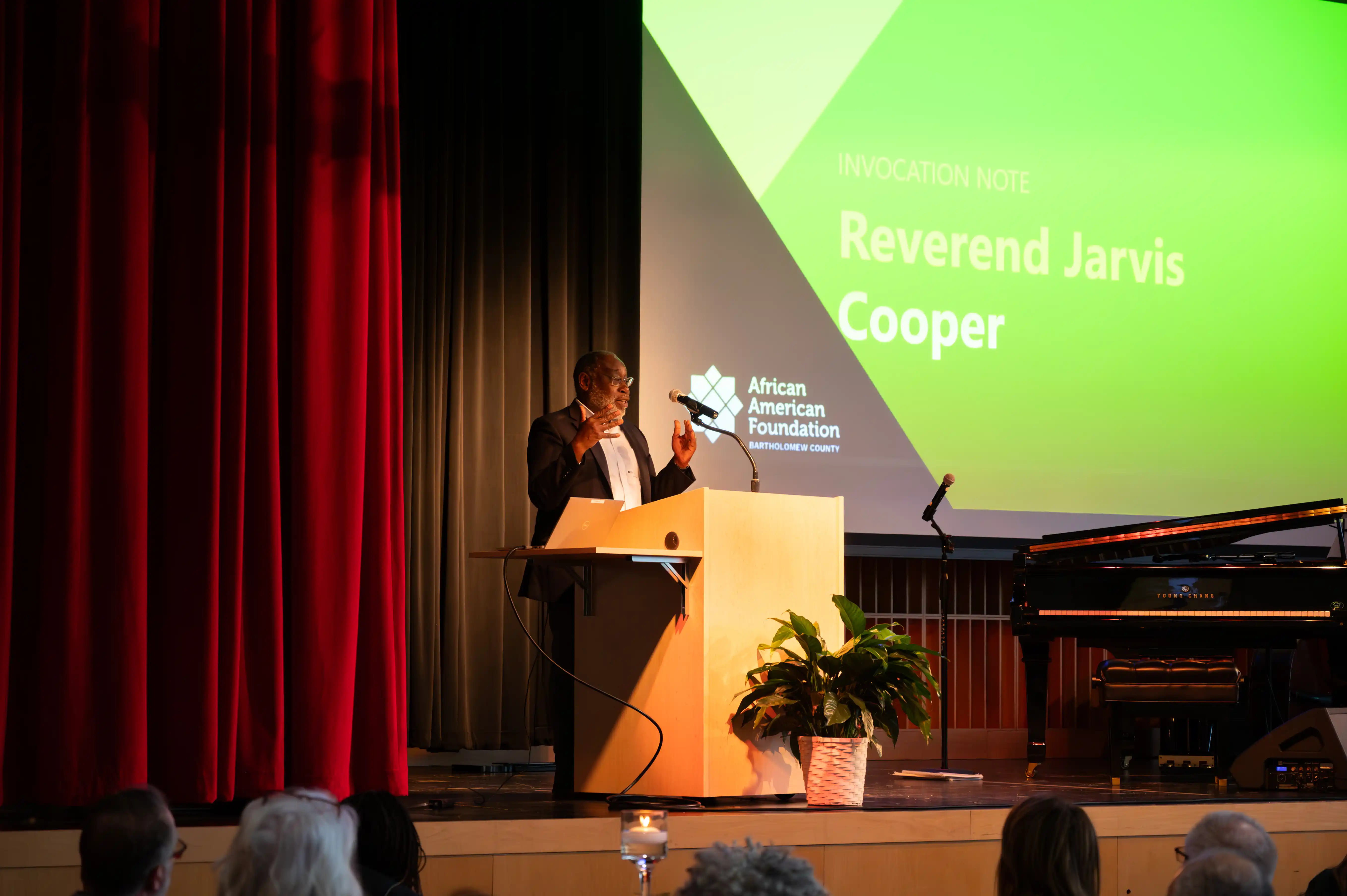 Person speaking at a podium with a projection screen in the background displaying "Reverend Jarvis Cooper" at an event.