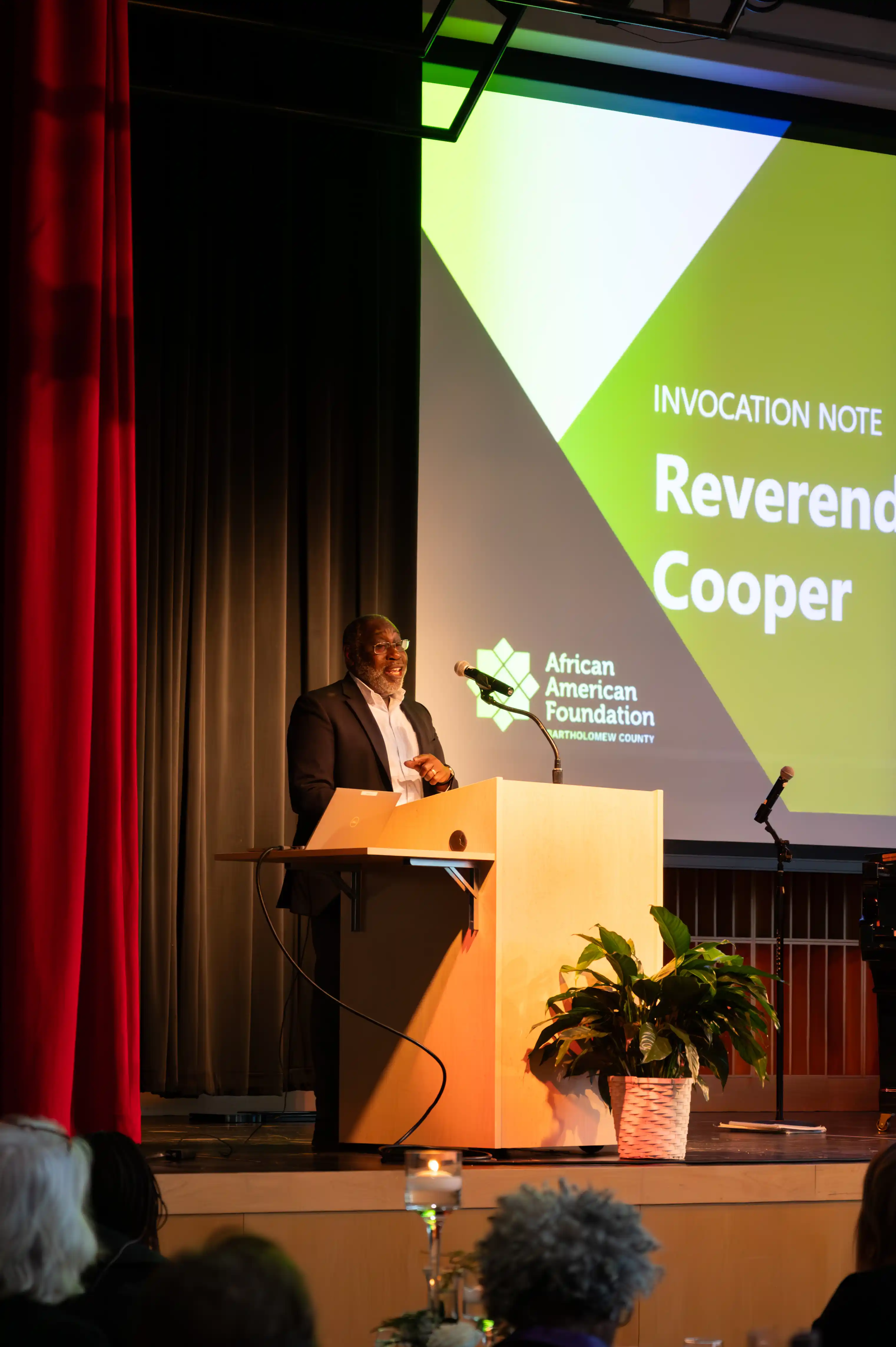 Speaker at podium presenting at a conference with a presentation screen in the background displaying the name Reverend Cooper.
