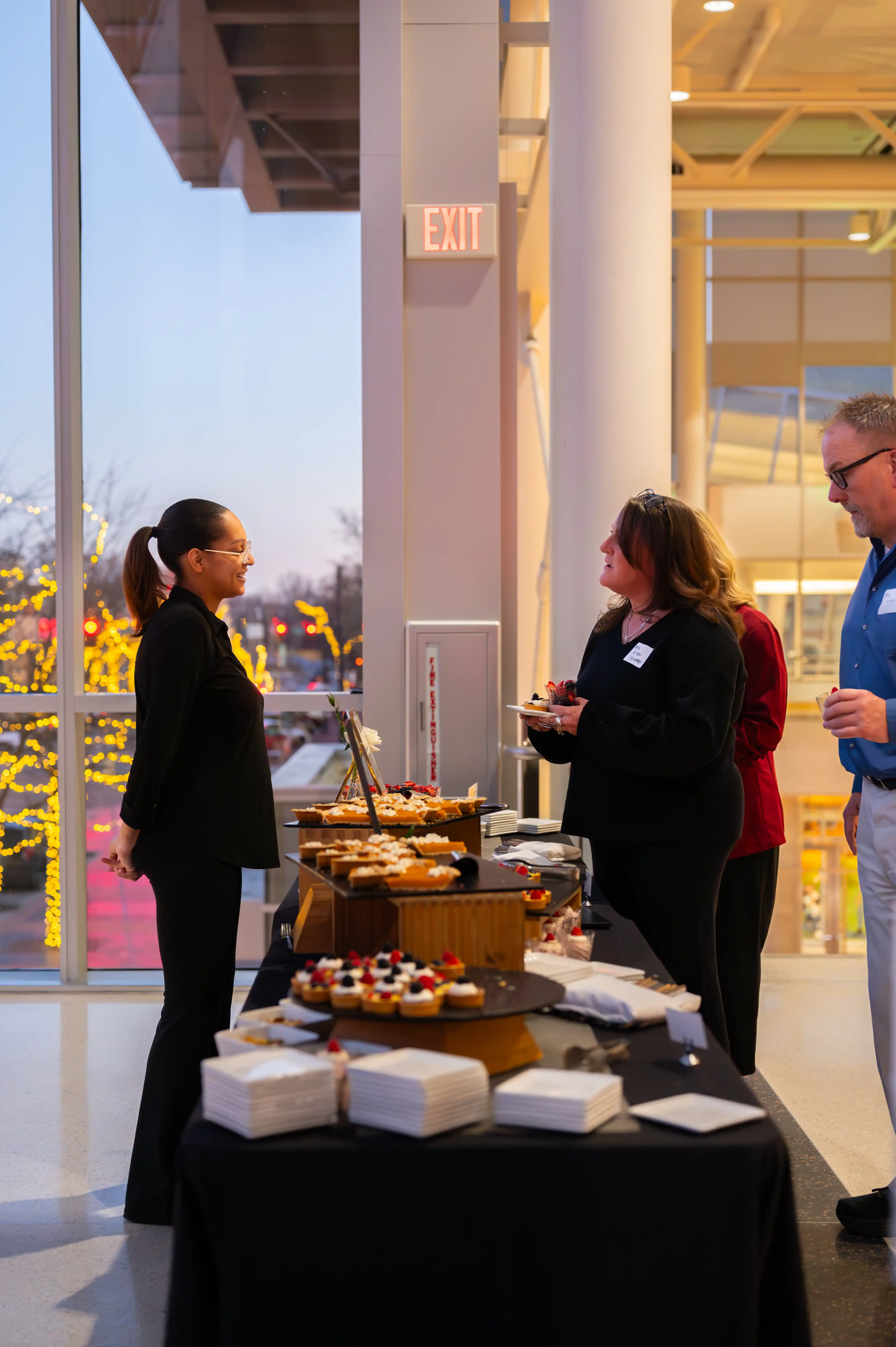 Two individuals conversing near a food table at an indoor event with windows showing a cityscape at dusk in the background.
