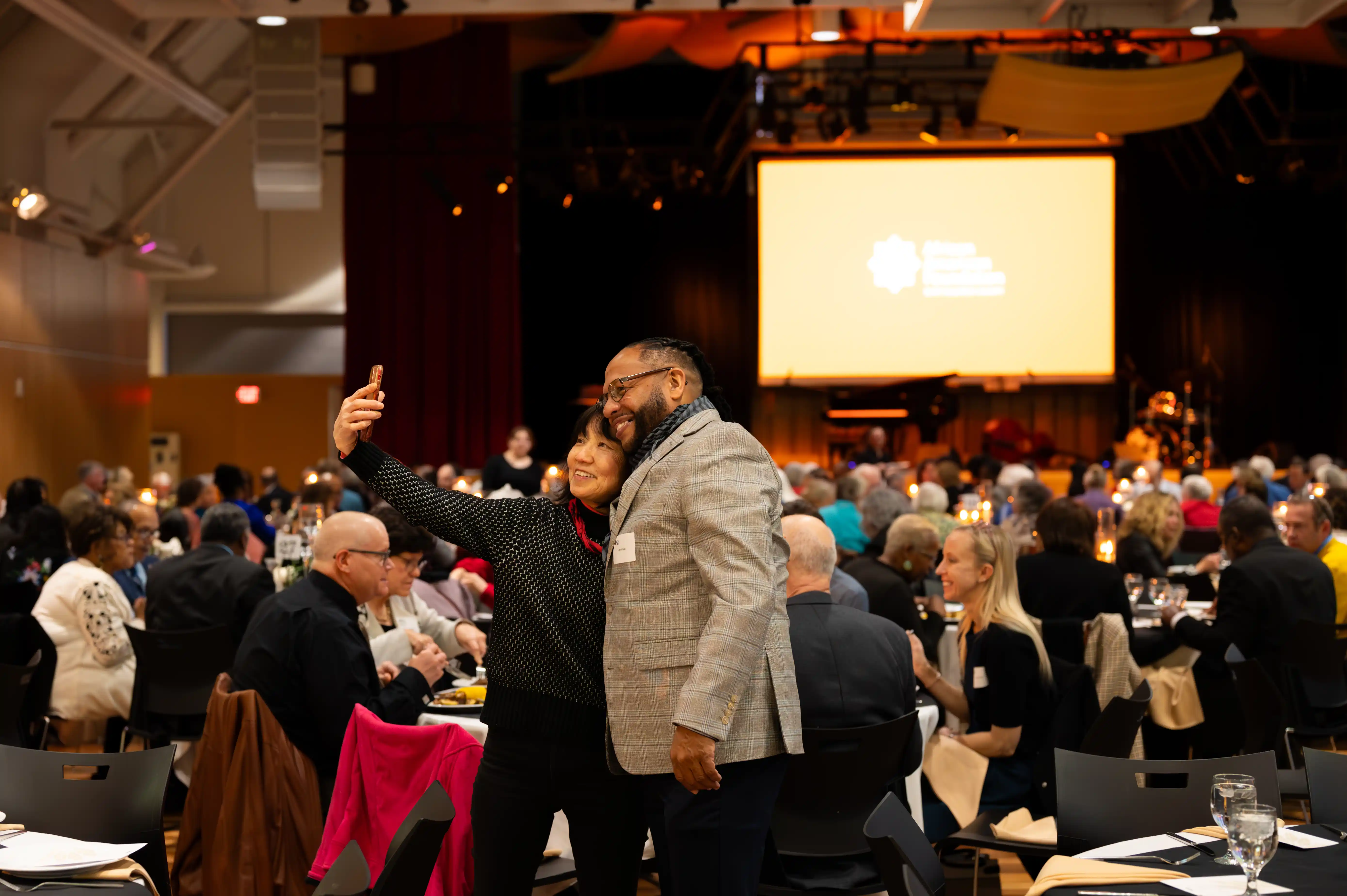 Two people taking a selfie in a bustling event hall with attendees seated at tables and a large screen in the background.