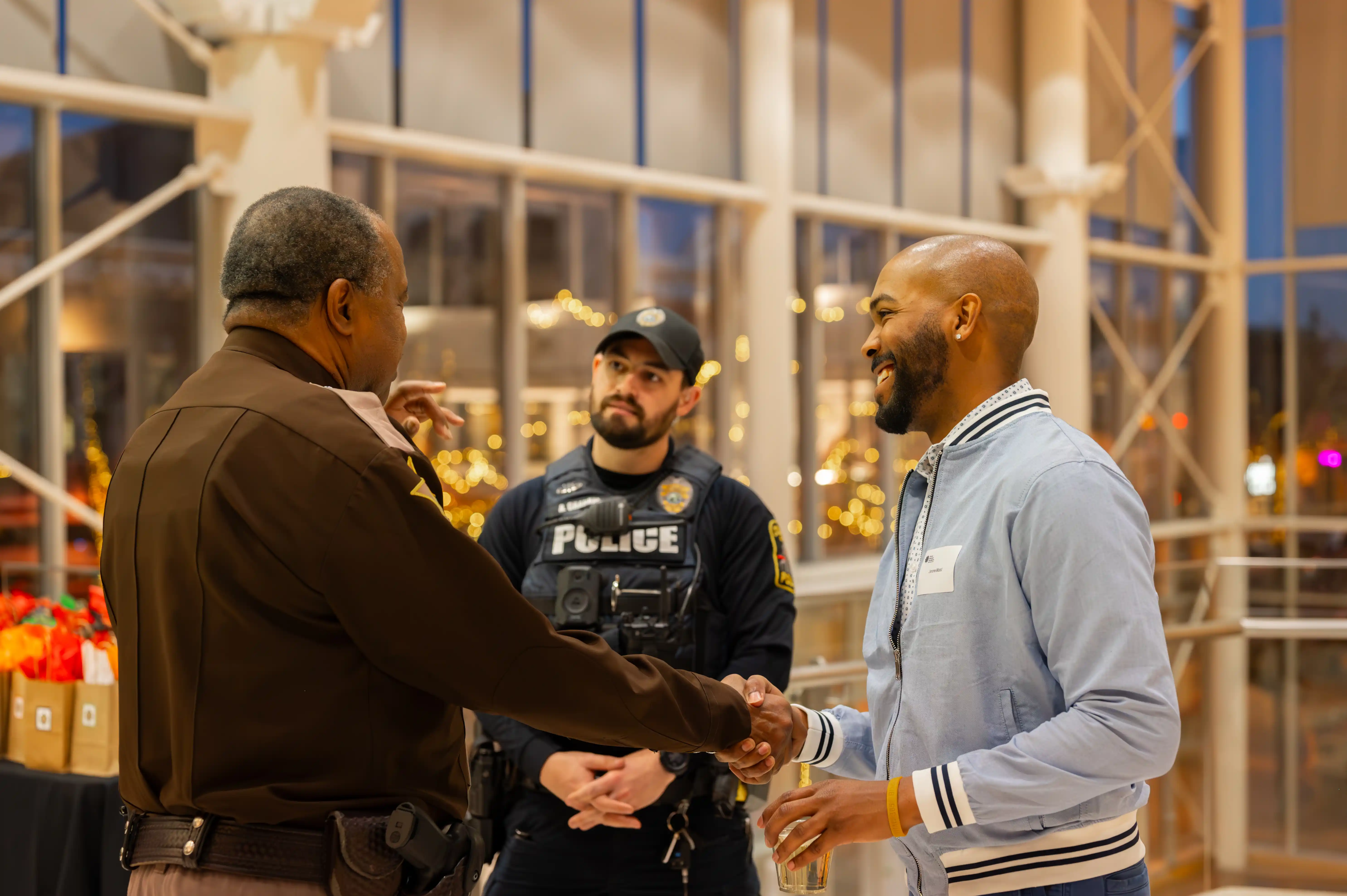 Two men shaking hands in a public indoor setting while another man in a police uniform observes.
