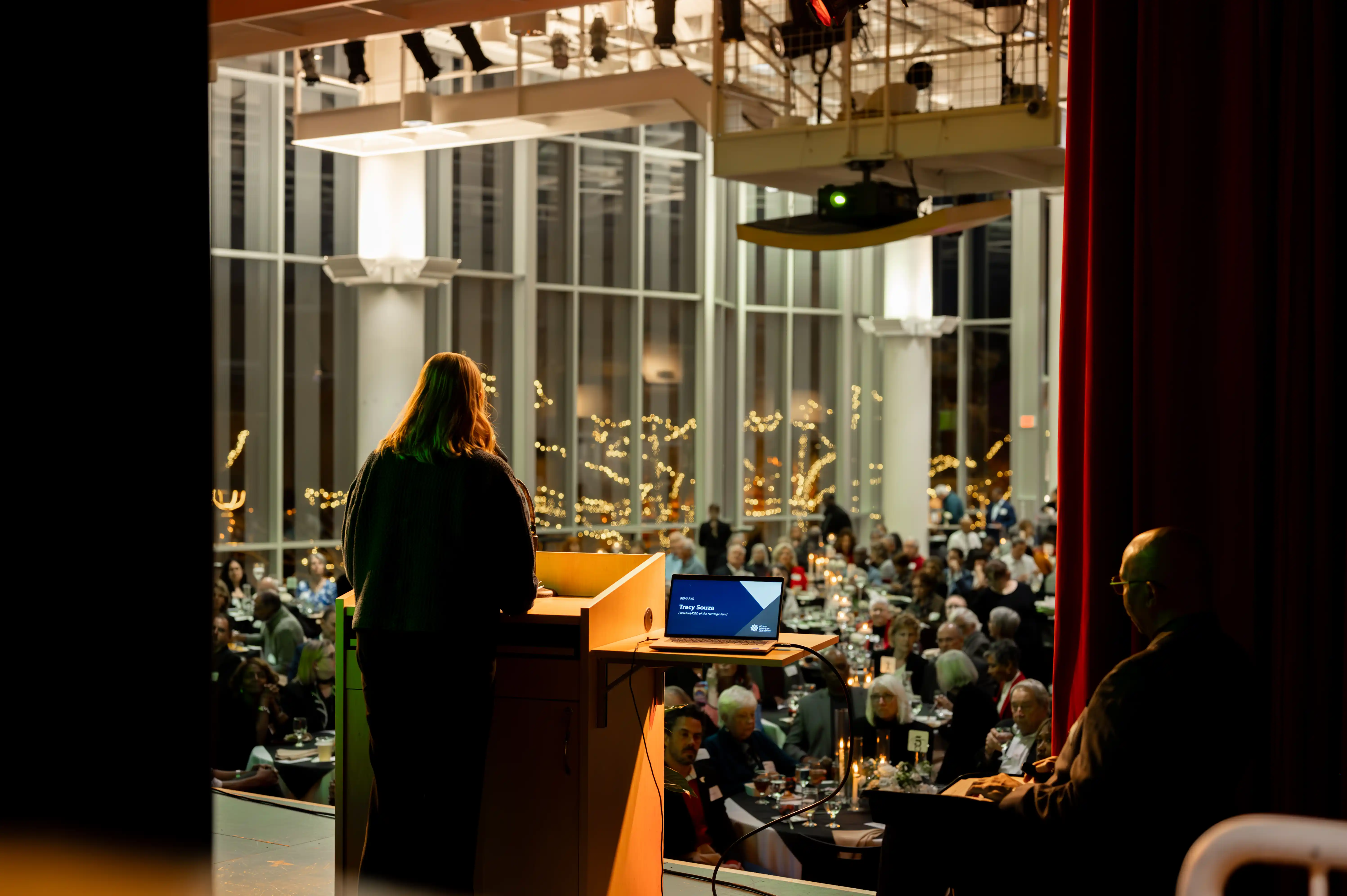 Person speaking at a podium in a banquet hall with guests seated at tables, viewed from behind the stage.