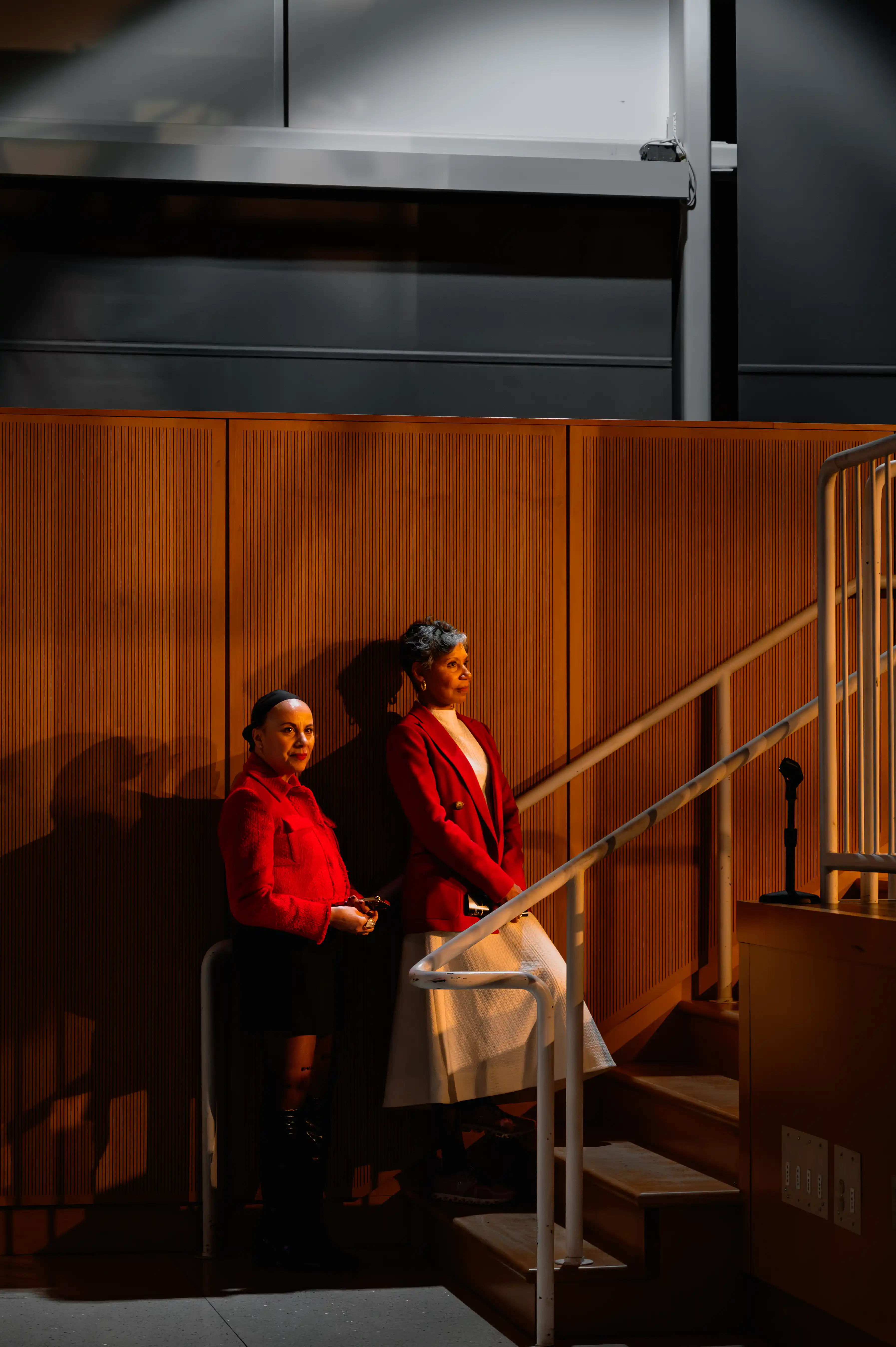 Two individuals in red tops seated on a staircase, with one holding a white handbag, in a dimly lit room with wooden walls.