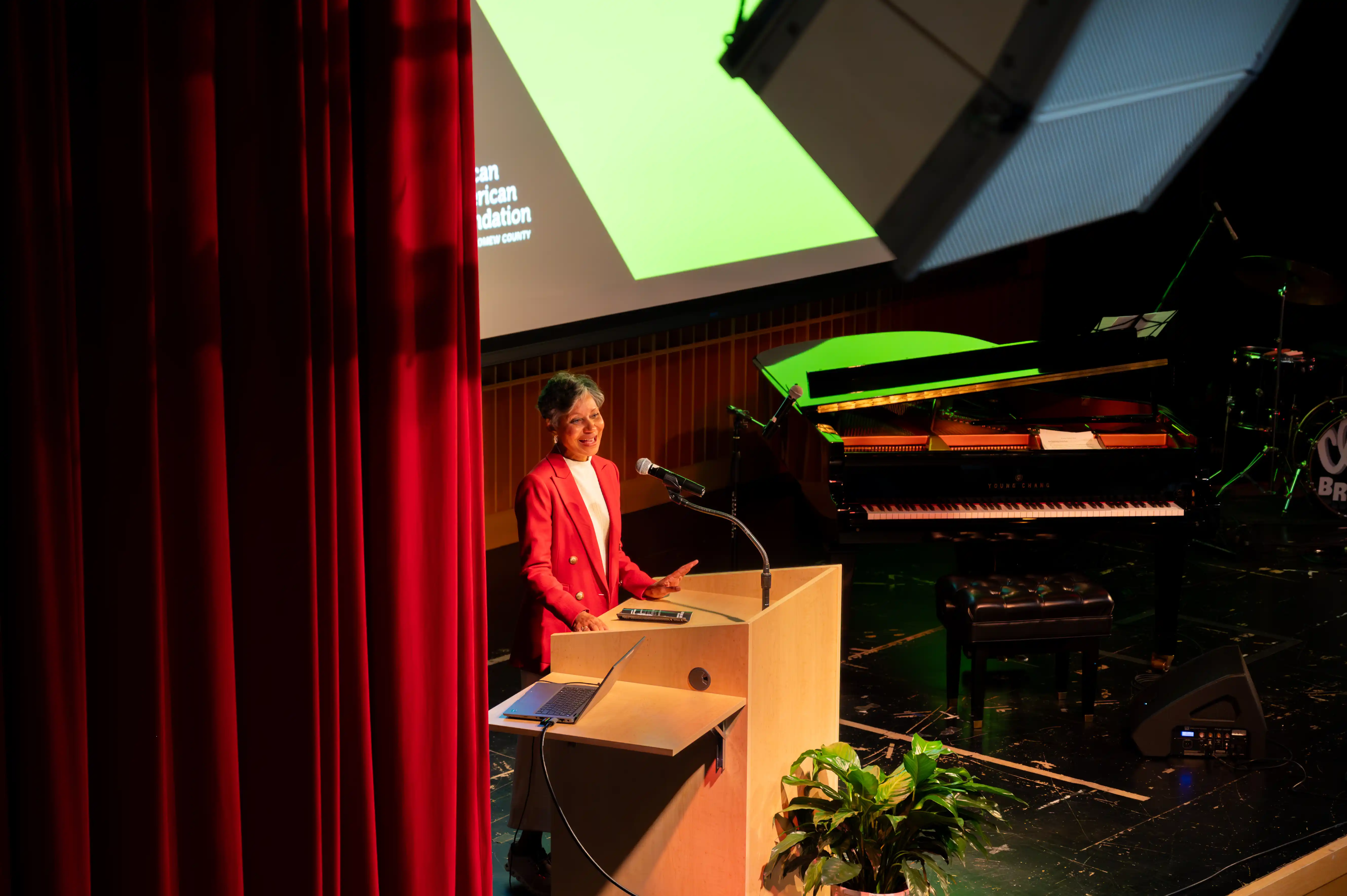 Woman speaking at a podium in an auditorium with a large screen in the background and a grand piano to the side.