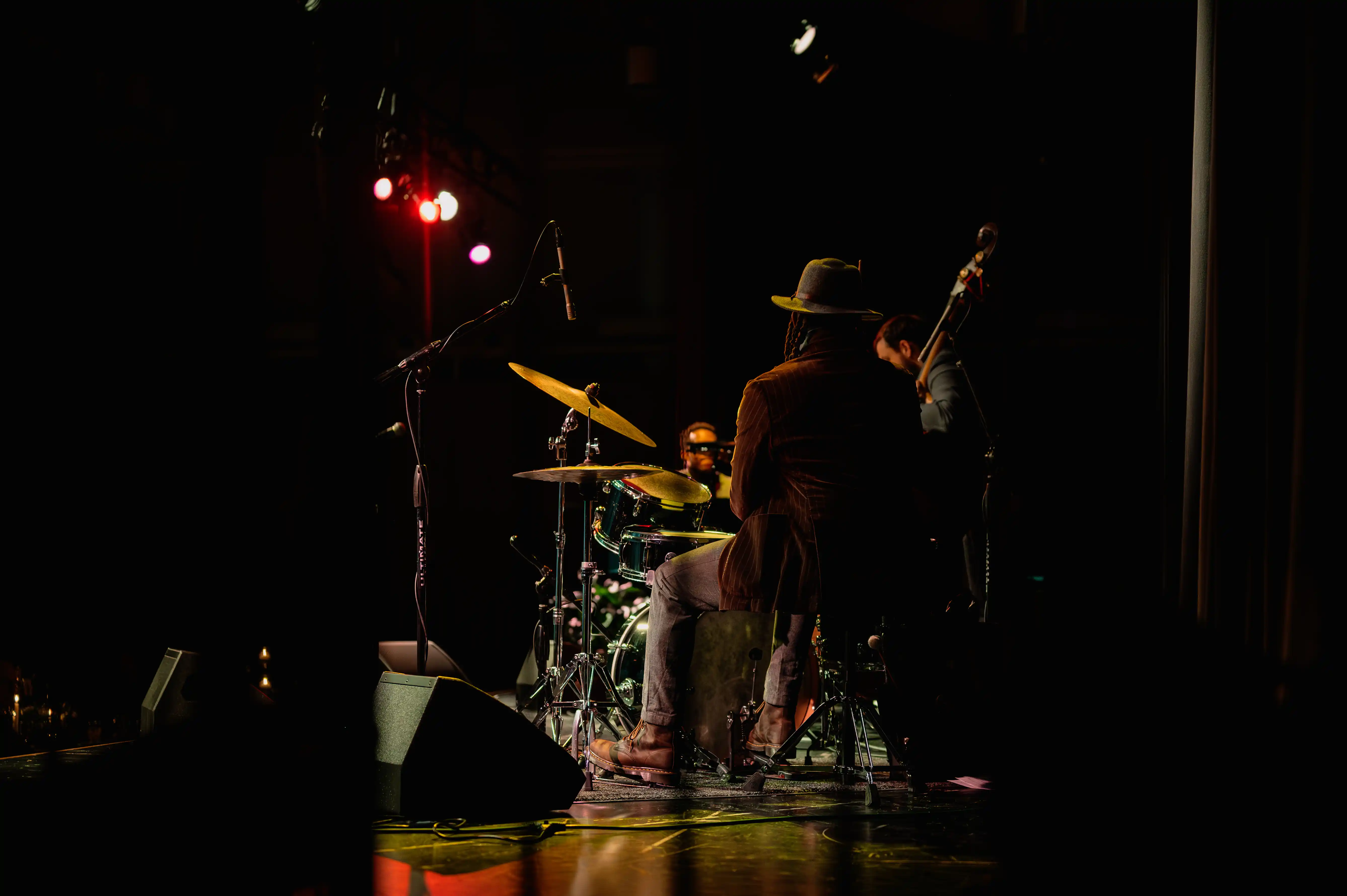 Musicians on stage with a drummer in the background and a person with a guitar in the foreground, dimly lit with red stage lighting.