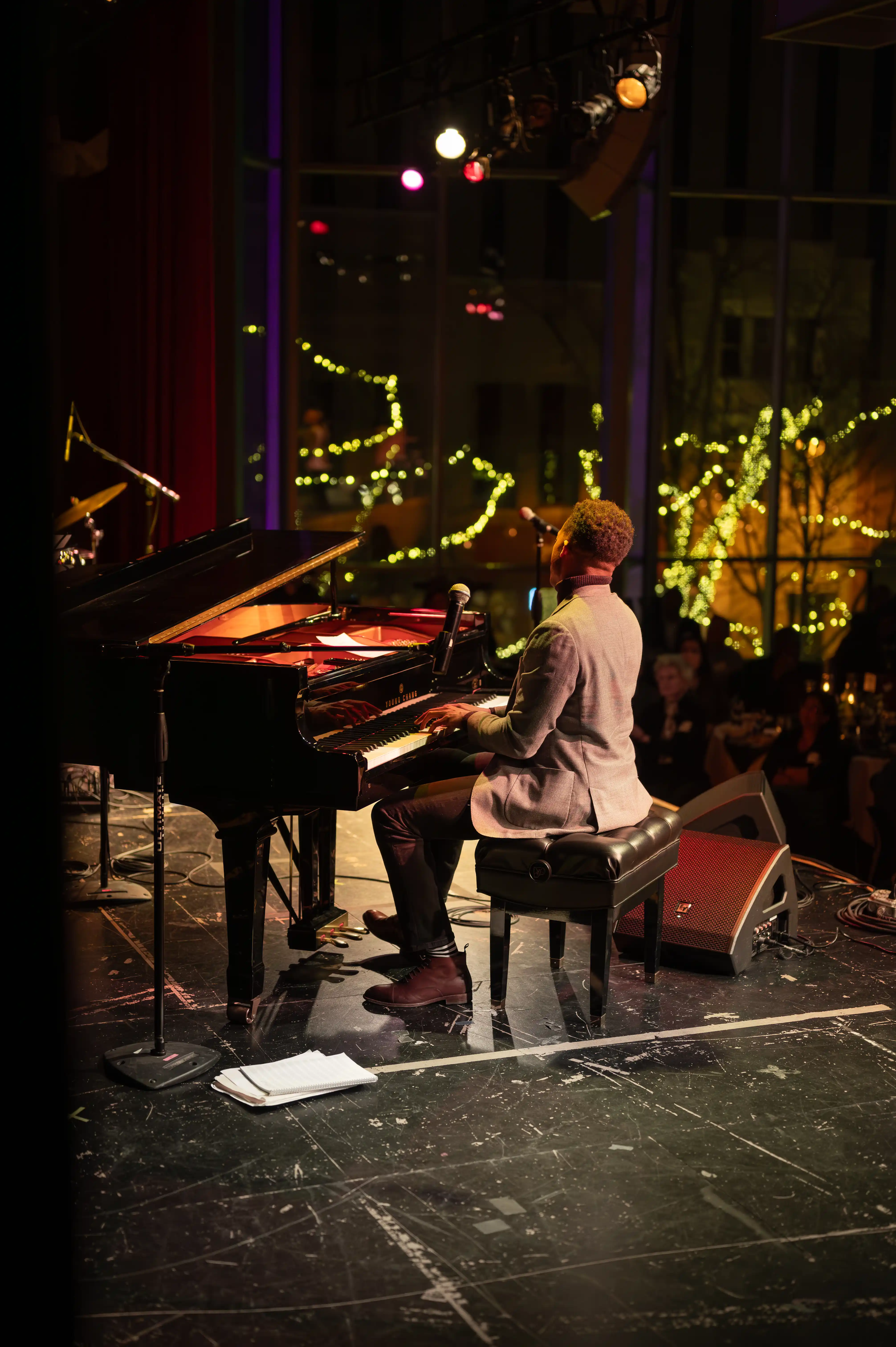 Pianist performing on stage with a grand piano in a dimly lit venue with soft lighting and reflections on the background windows.