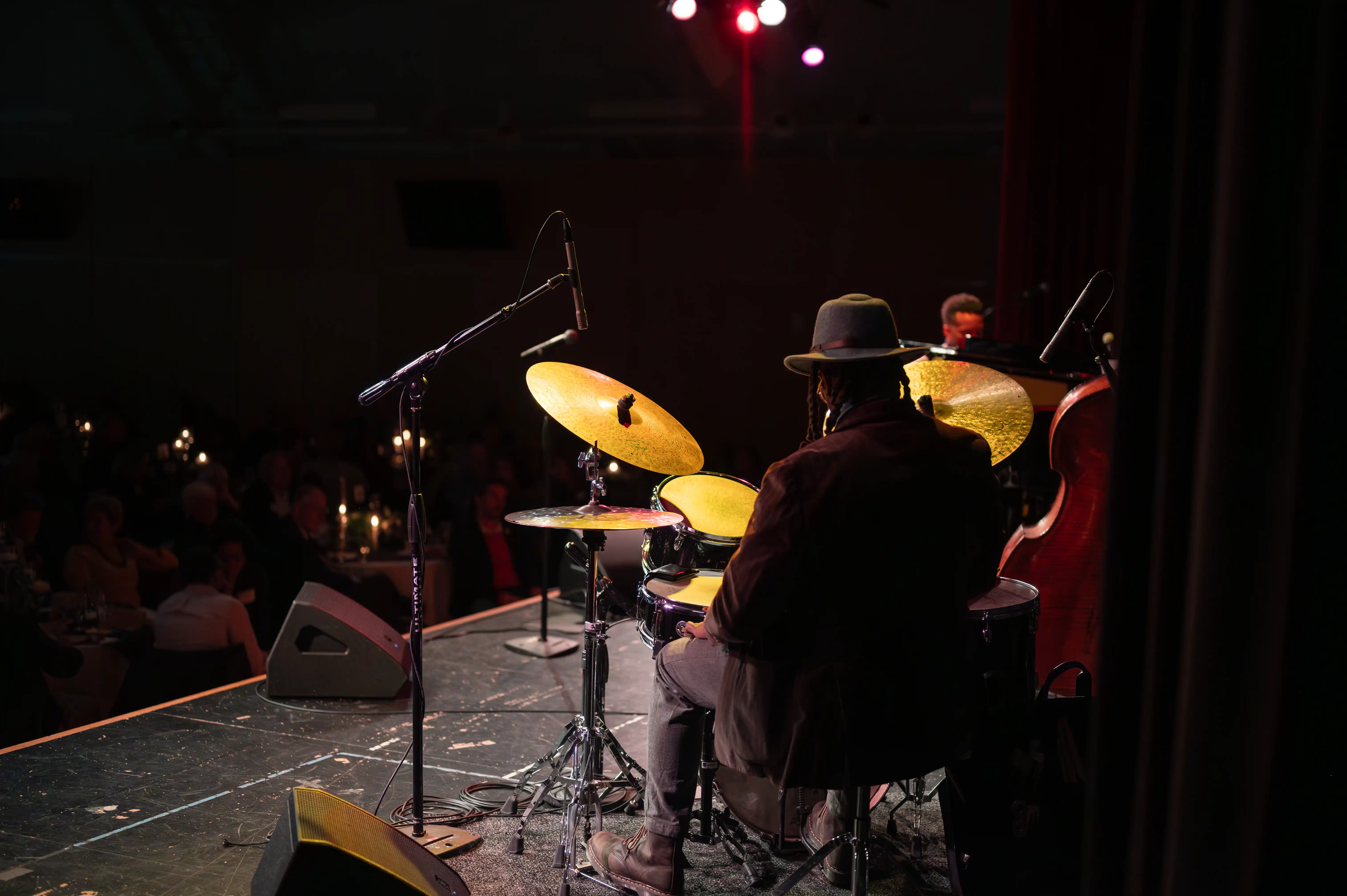 Jazz musicians performing on stage, drummer in focus, with a double bass player in the background under red stage lights.