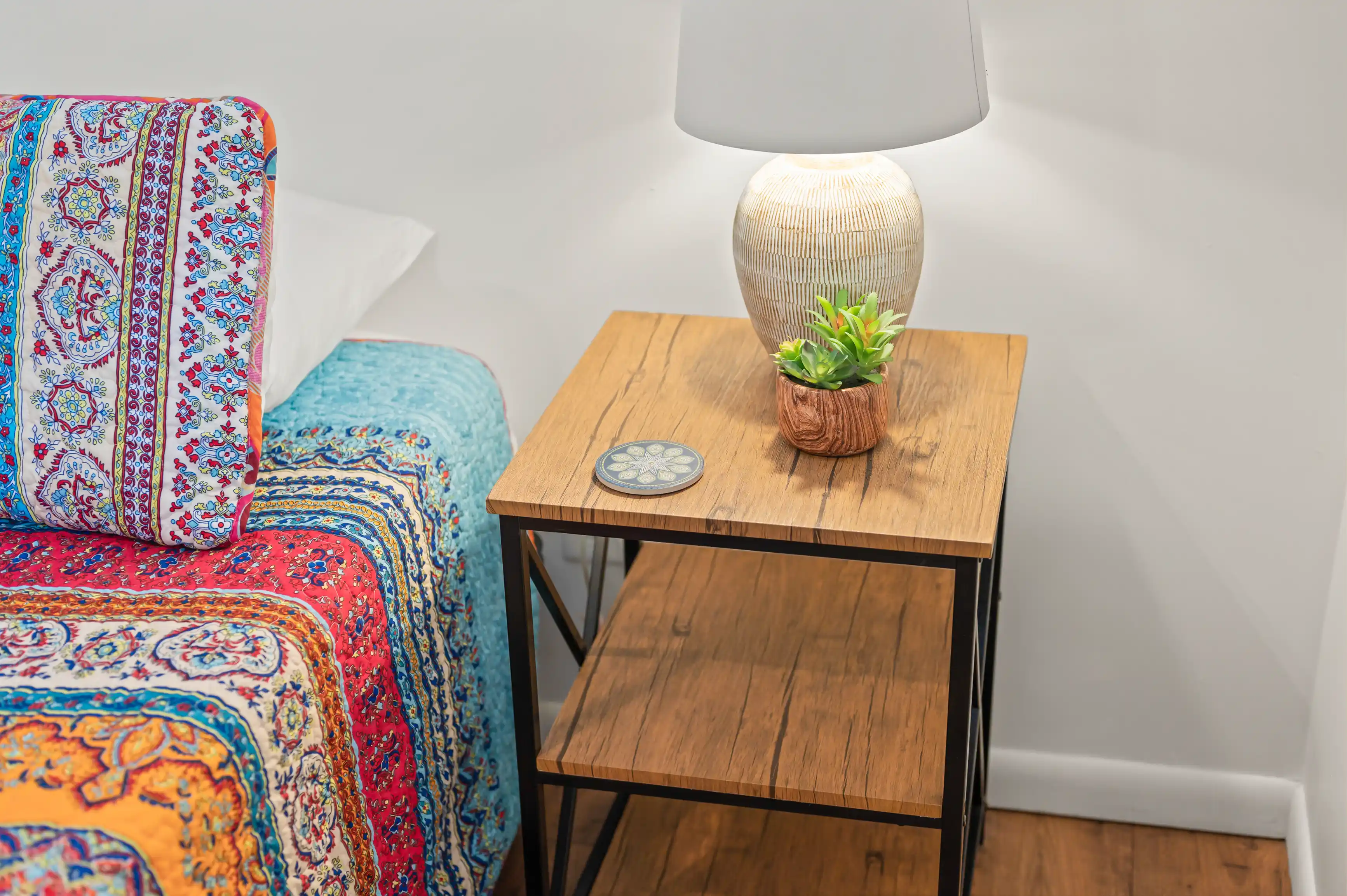 Cozy corner with a colorful patterned bedspread and a nightstand featuring a ceramic lamp and a small potted plant.