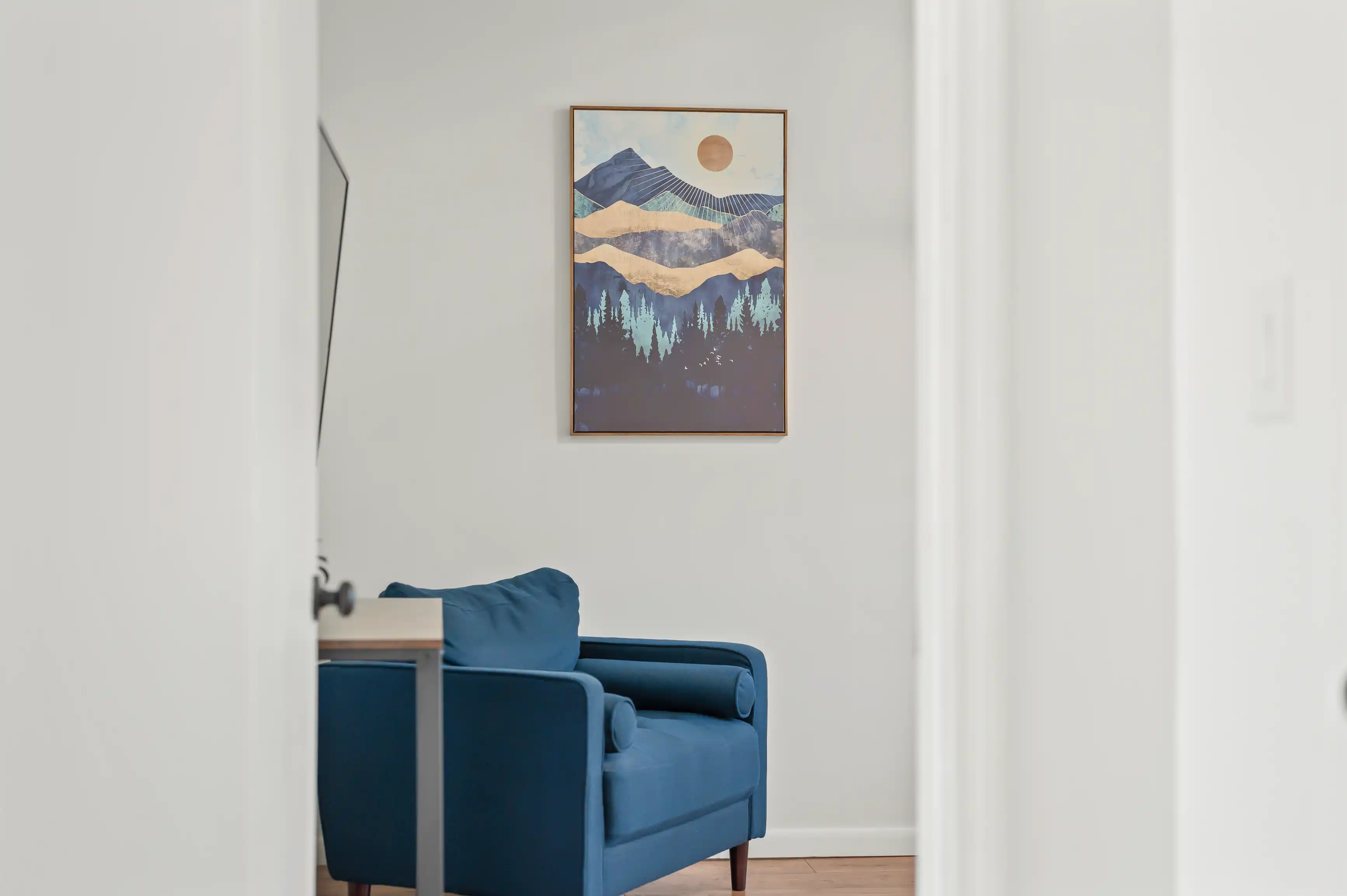 A minimalist living room corner with a blue armchair and framed mountainous landscape artwork on the wall.