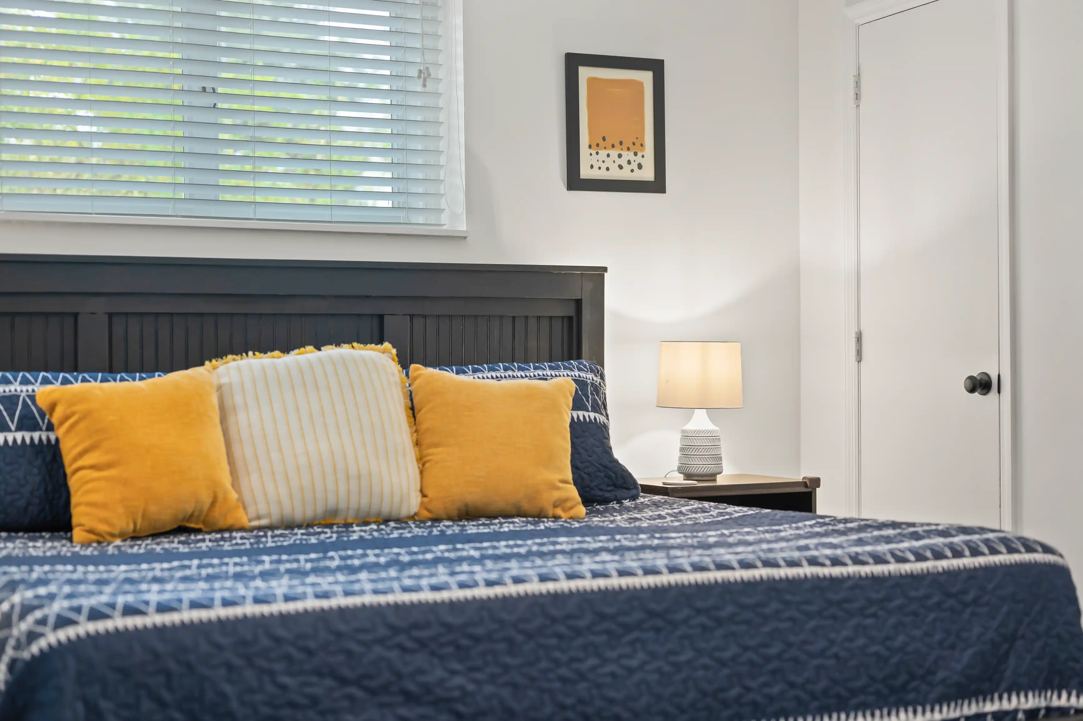 A cozy bedroom with a neatly made bed adorned with blue and yellow pillows, a lamp on a bedside table, closed white blinds on the window, and a framed artwork on the wall.