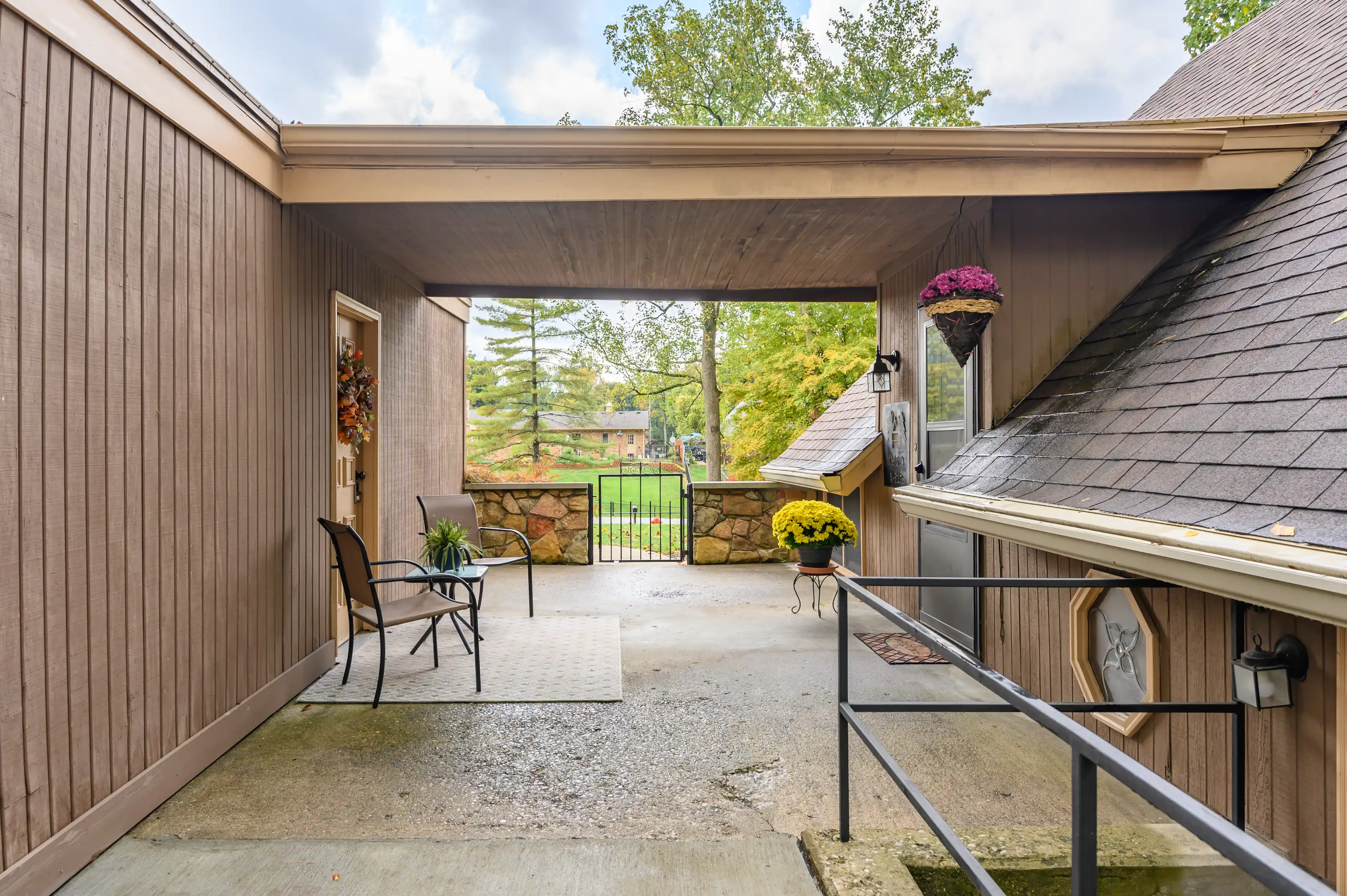 Covered carport area with a metal gate leading to a green lawn, adorned with hanging and potted flowers, and outdoor seating by the house door.