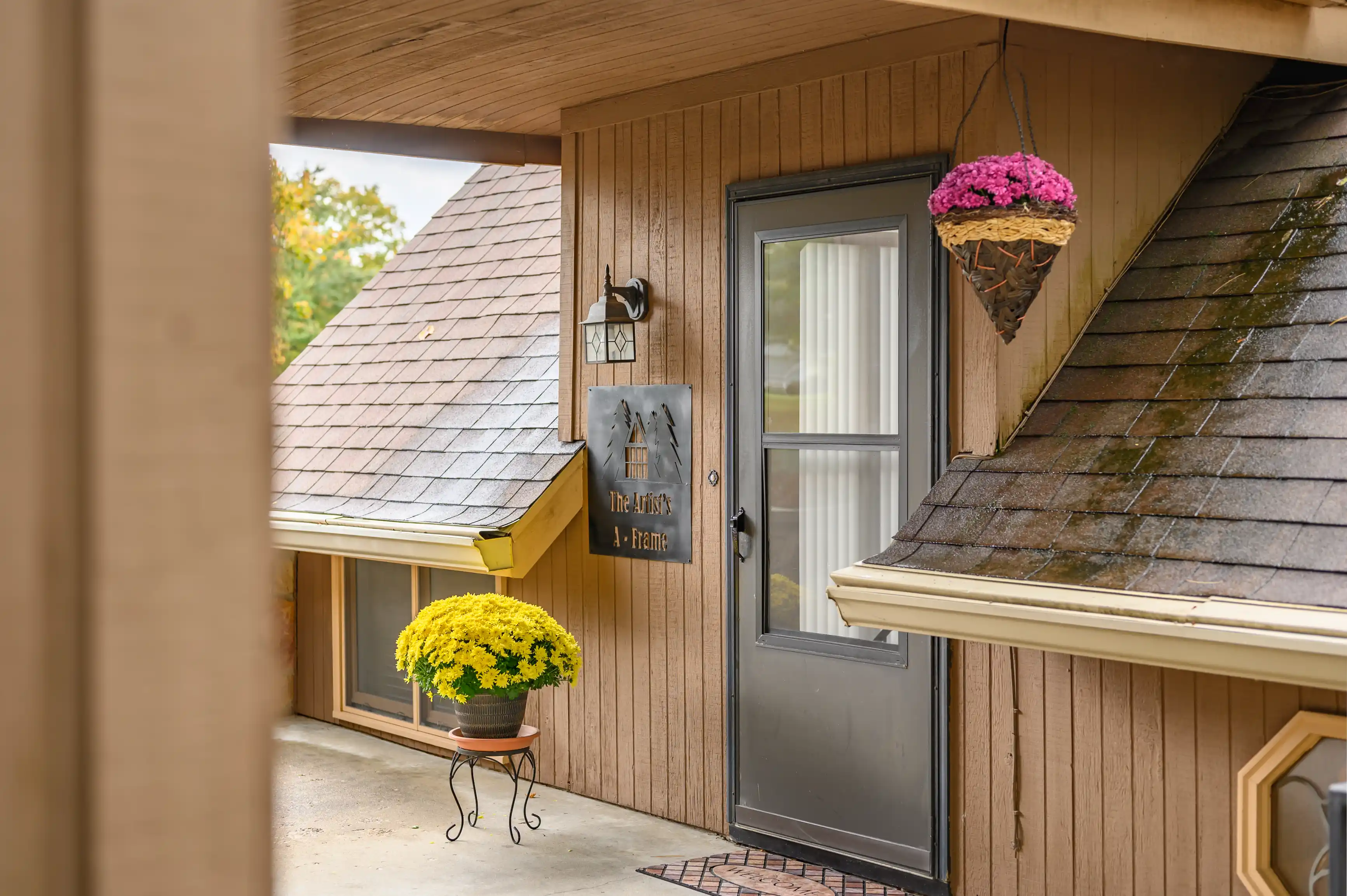 A cozy house entrance with a wooden front door, a hanging flower basket, and a potted yellow chrysanthemum by the doorstep, with a sign reading "The Artist's A-Frame".