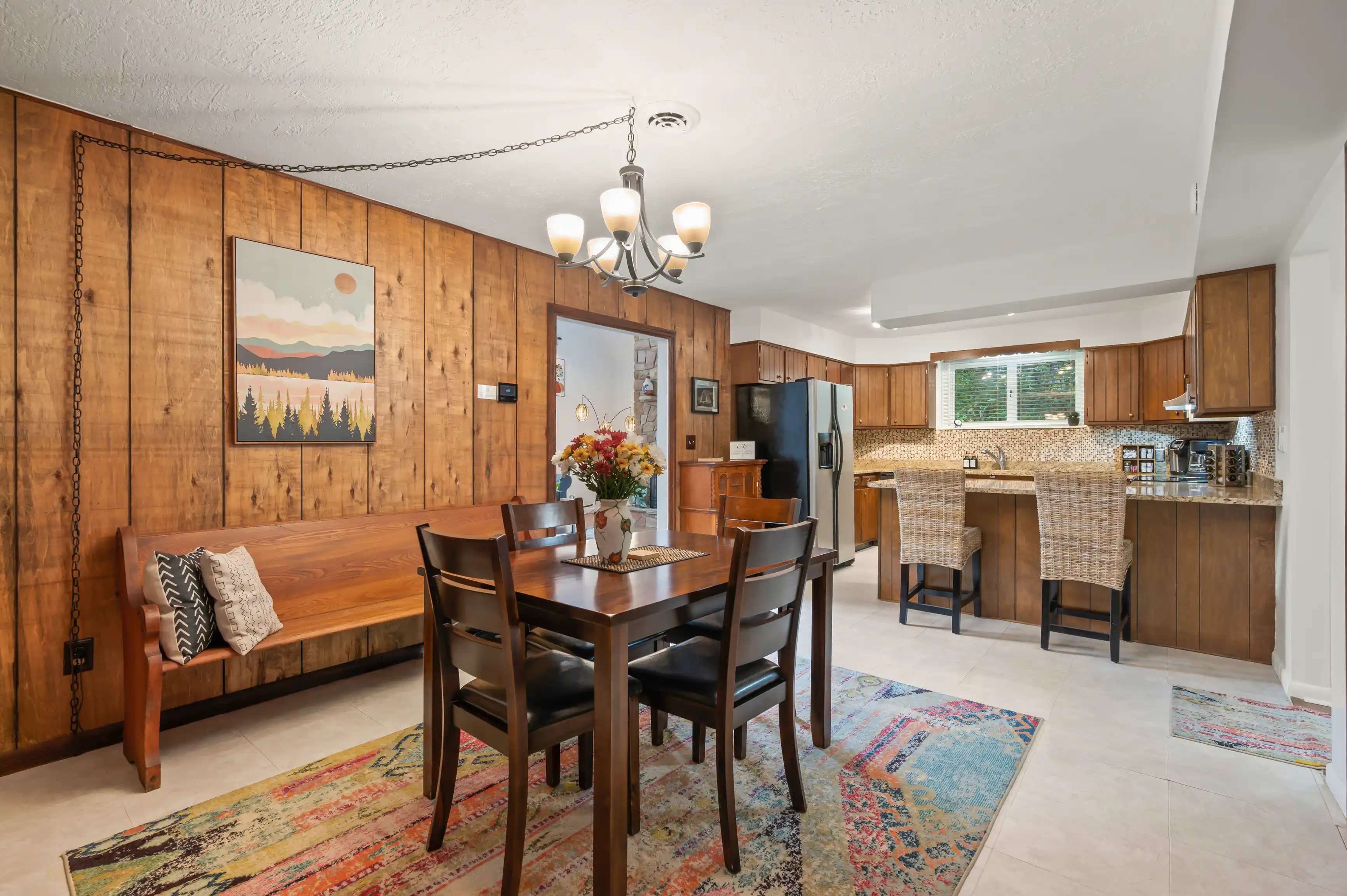 A cozy dining area with a wooden table and chairs on a colorful rug, adjacent to a kitchen with wooden cabinetry and modern appliances. A scenic landscape painting adorns the wood-paneled wall.