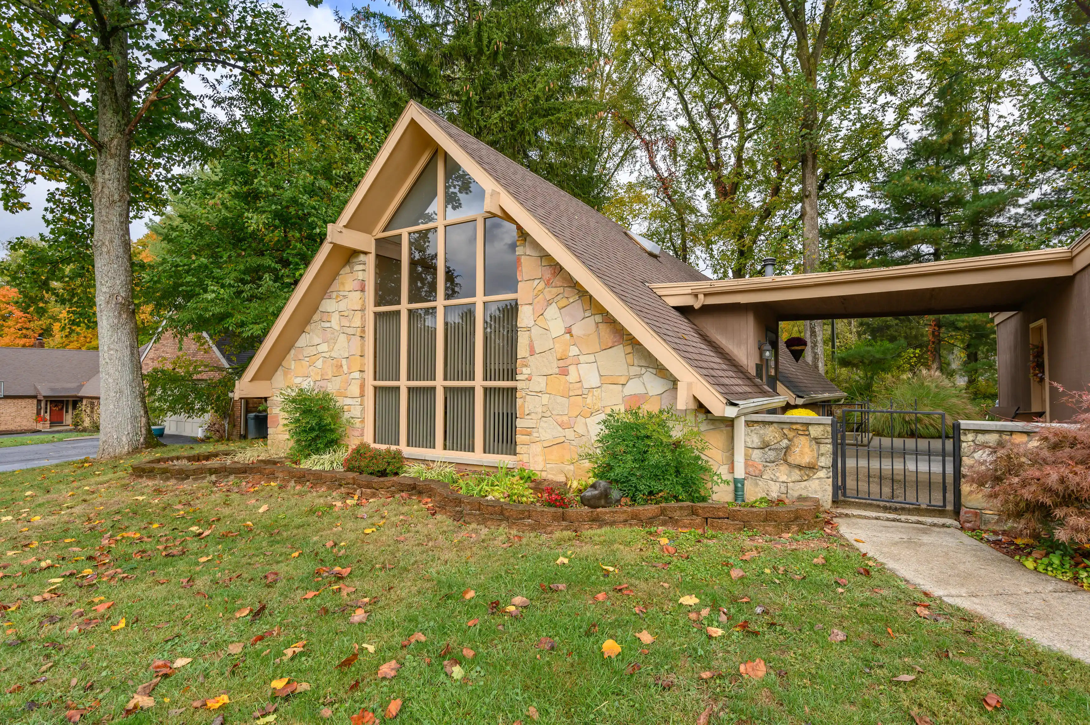 A unique A-frame house with large windows, stone accents, and a carport surrounded by trees in an autumn setting.
