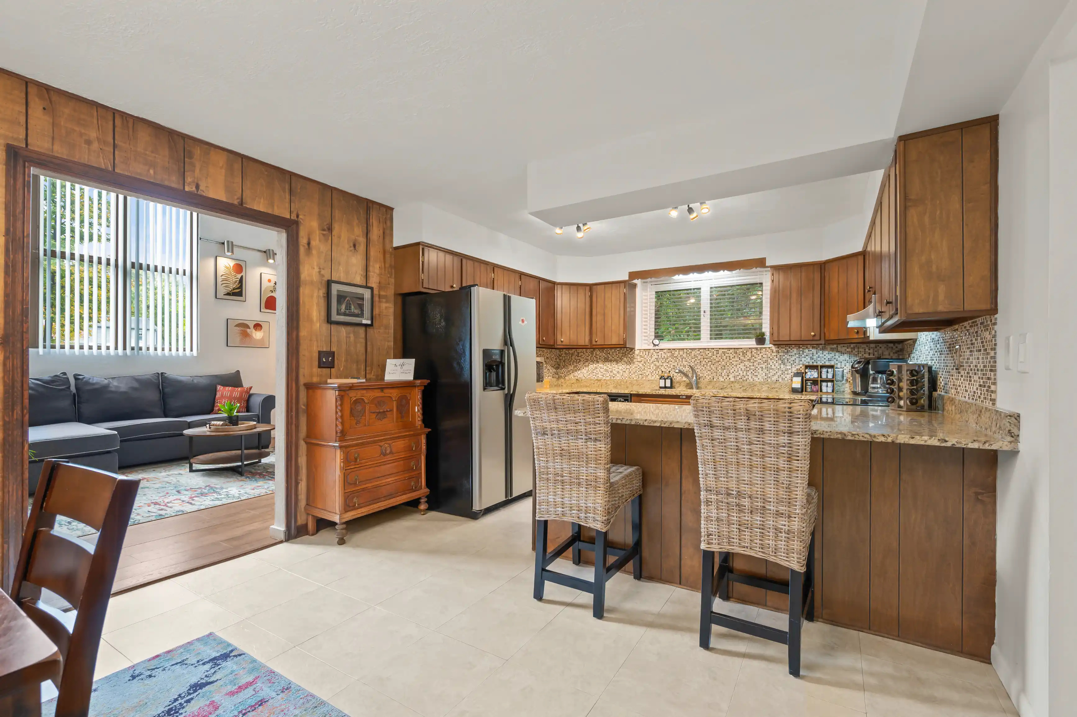Spacious kitchen with wooden cabinets and modern appliances, featuring a tiled backsplash and a bar seating area with wicker stools. Adjacent area reveals a cozy living room with a dark leather sofa and decorative art on the walls.