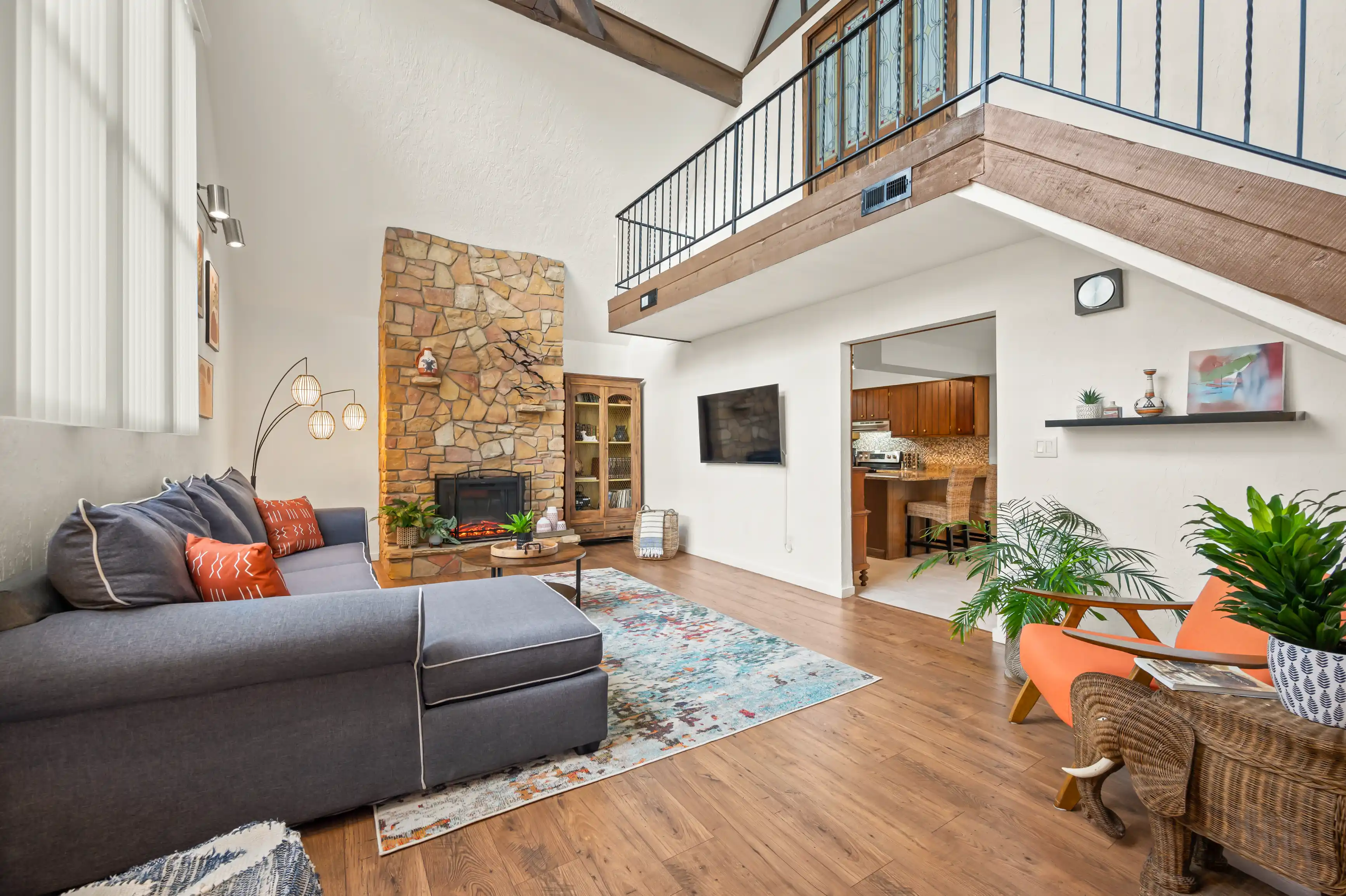 Spacious living room with high ceiling, stone fireplace, L-shaped sofa, colorful rug, wooden flooring, and an upper level balcony with railing.