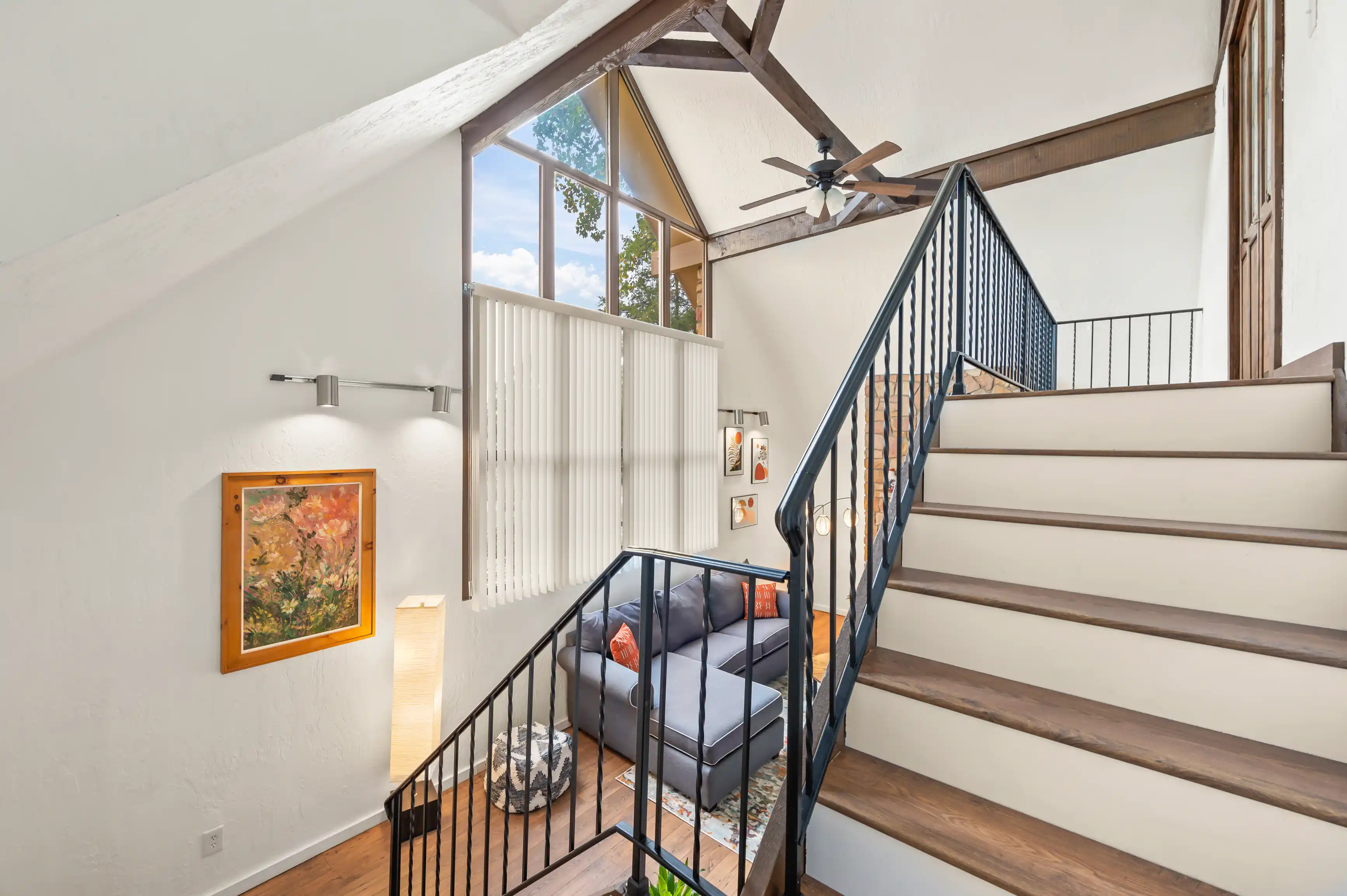 Bright, modern stairwell with large windows, wooden steps, a metal railing, a ceiling fan, and a cozy sitting area with a couch and artwork.