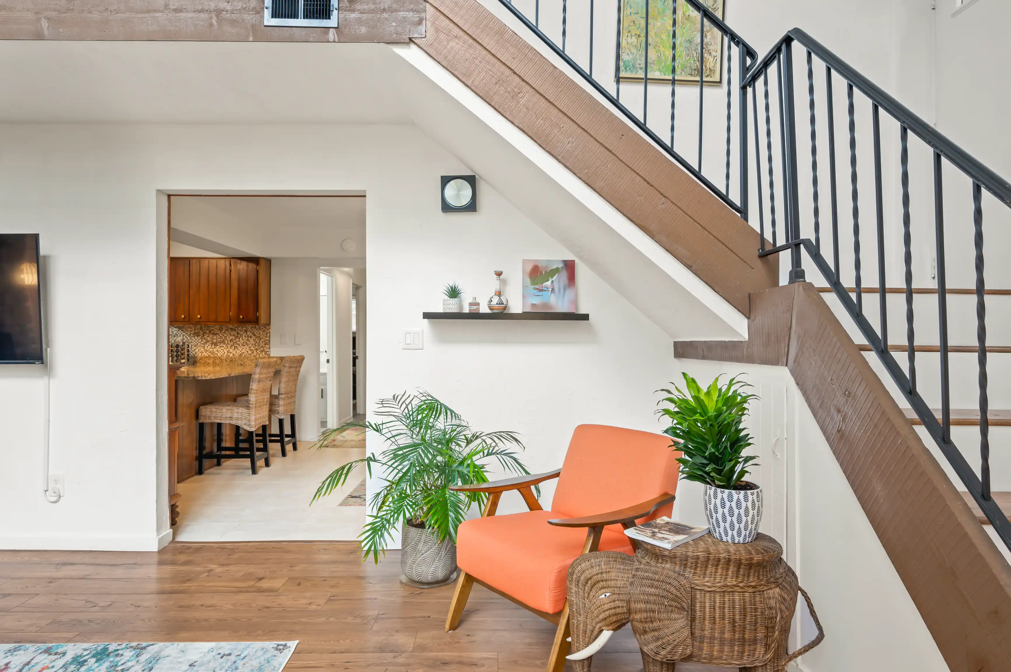 Bright, modern living room interior with orange chair, wicker furniture, indoor plants, wooden staircase with metal banisters, and a kitchen in the background.