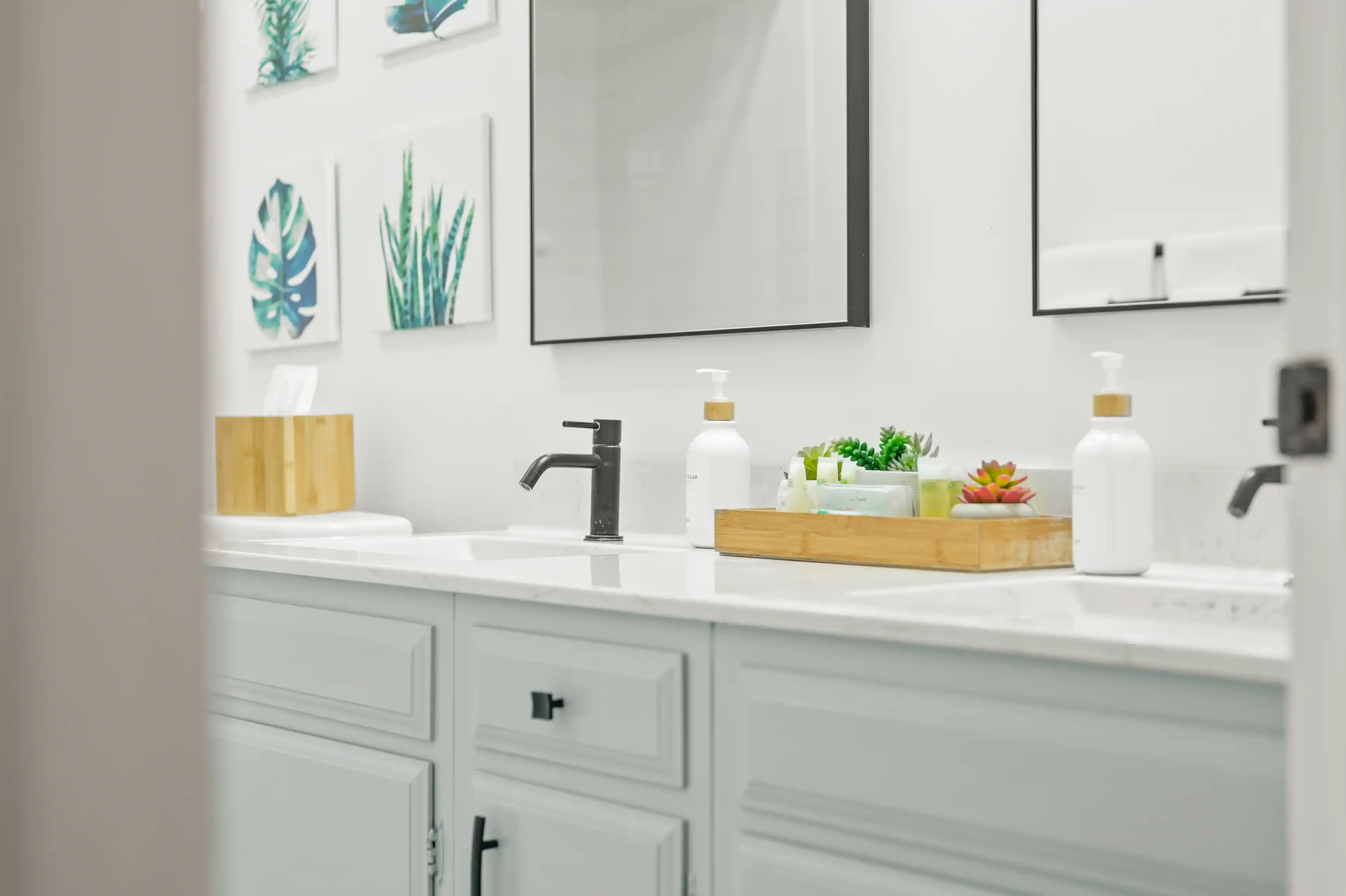 A modern bathroom vanity with a white countertop, black faucet, framed mirrors, and decorative plants.
