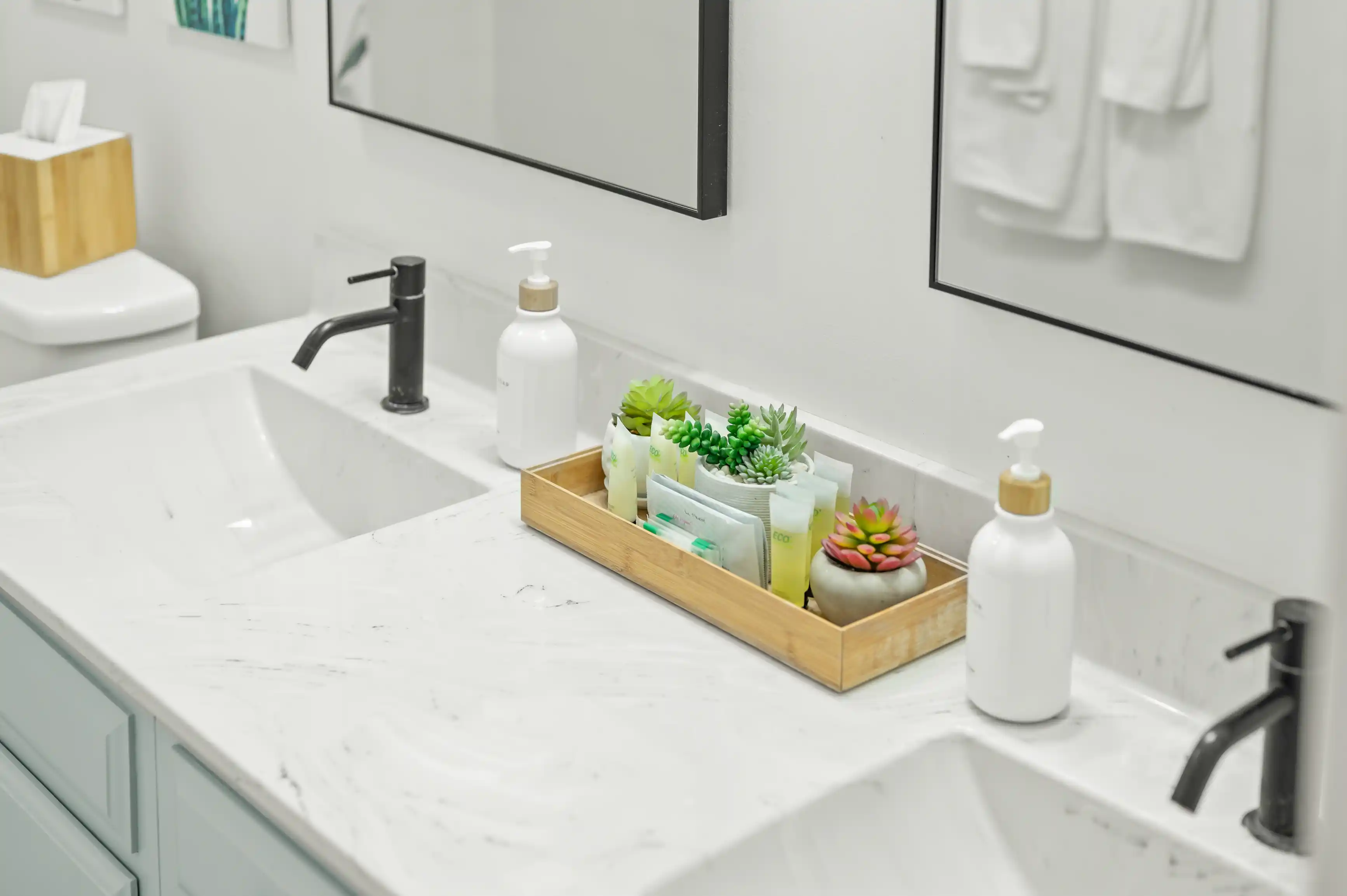 Modern bathroom interior with marble countertop and sink, black faucet, and various toiletries and plants arranged in a wooden tray.