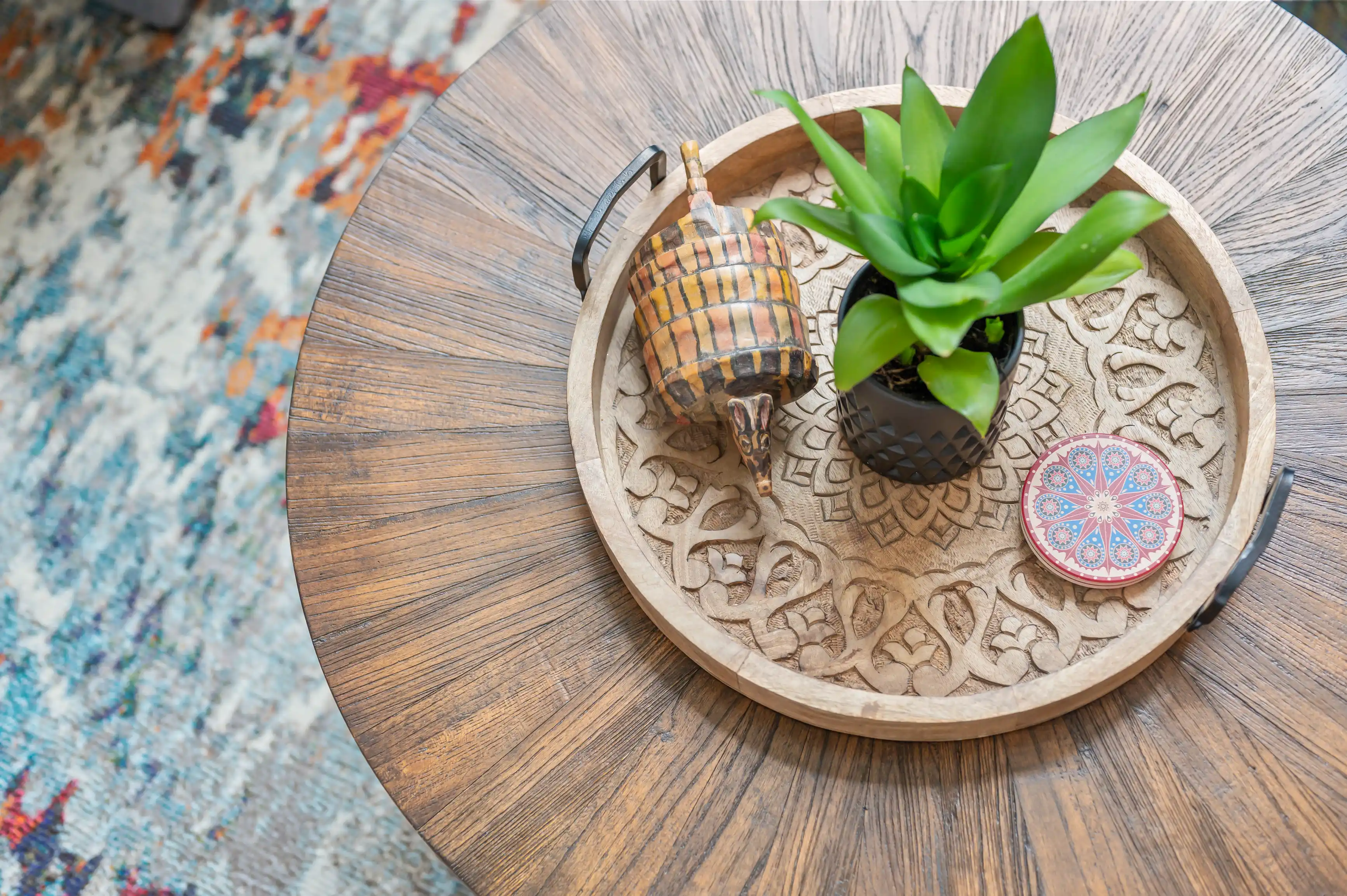Decorative round wooden tray with intricate carving containing a succulent plant in a geometric pot, a small checkered decorative armadillo, and a red patterned coaster on a wooden floor with a colorful rug edge visible.