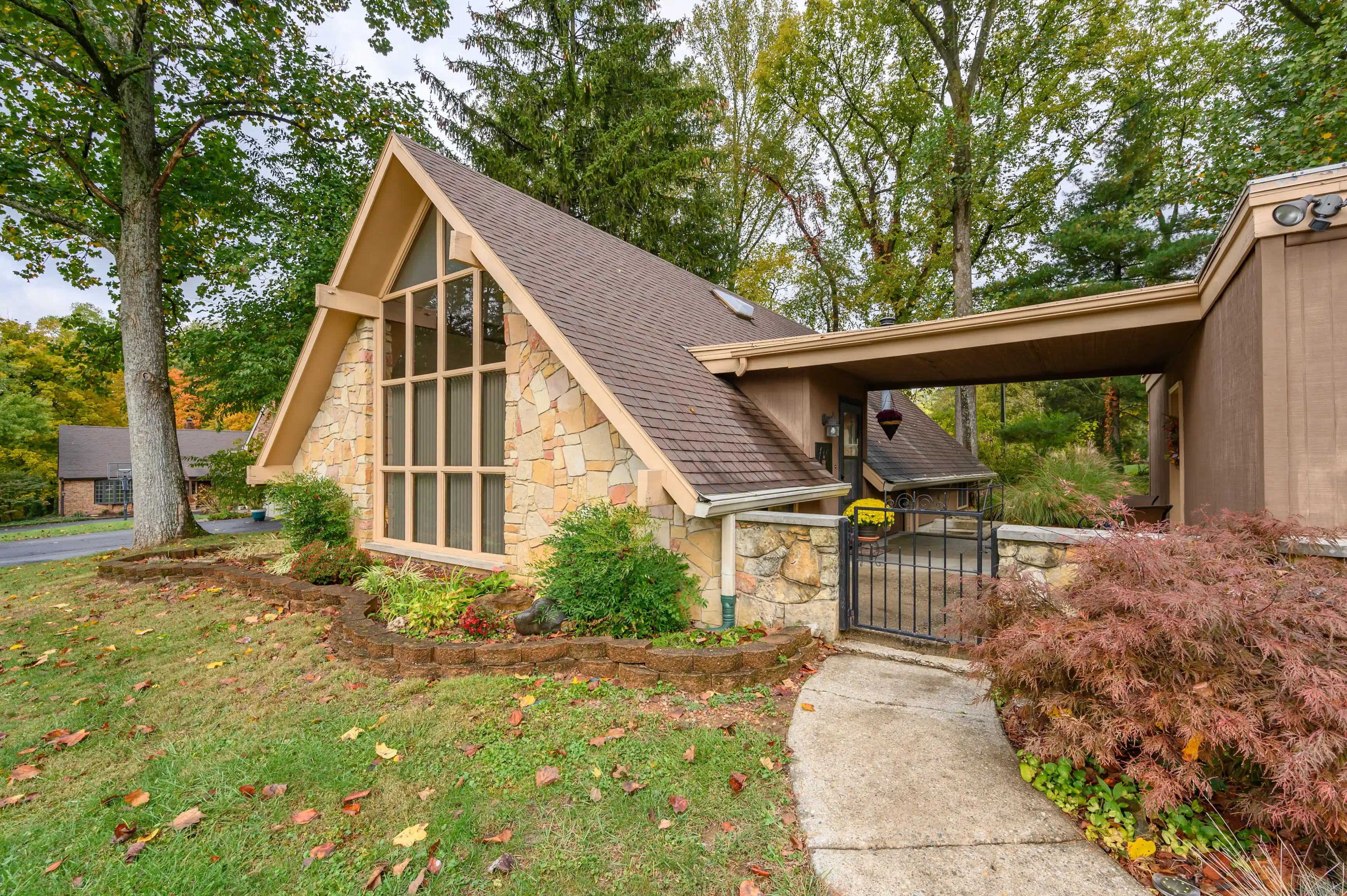 Unique A-frame house with large windows surrounded by autumn foliage, featuring a stone facade and an attached carport.